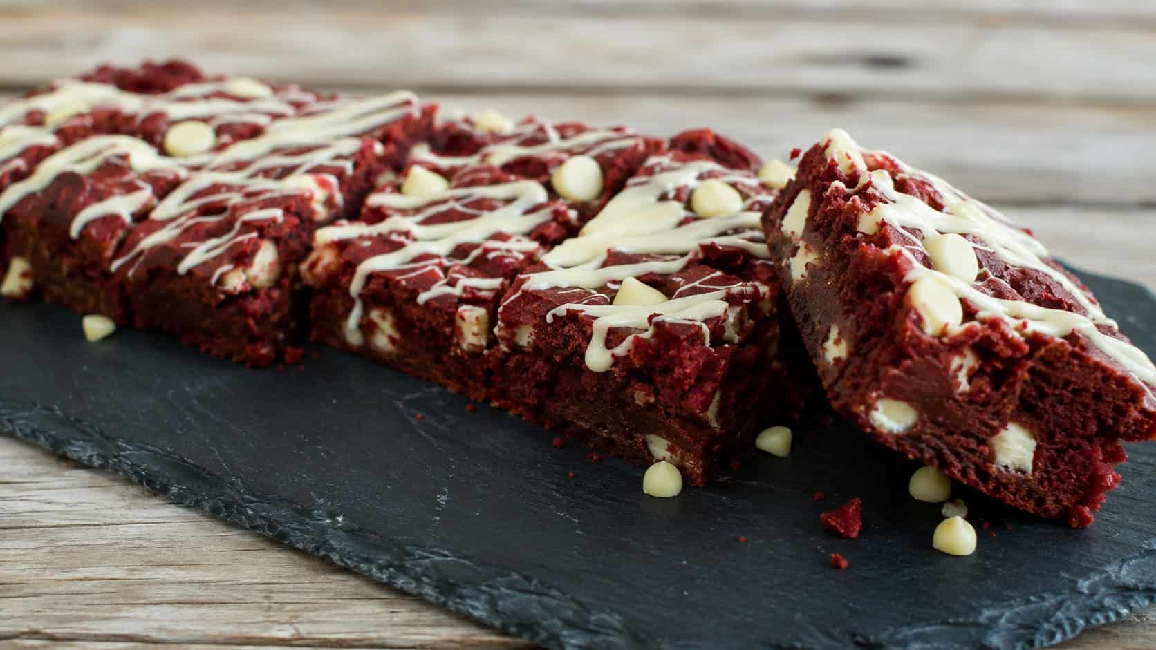 a wide horizontal arrangement of deep red brownies with creamy white chocolate chips and a white chocolate drizzle on top, on a dark slate board, natural light, shallow depth of field, landscape orientation