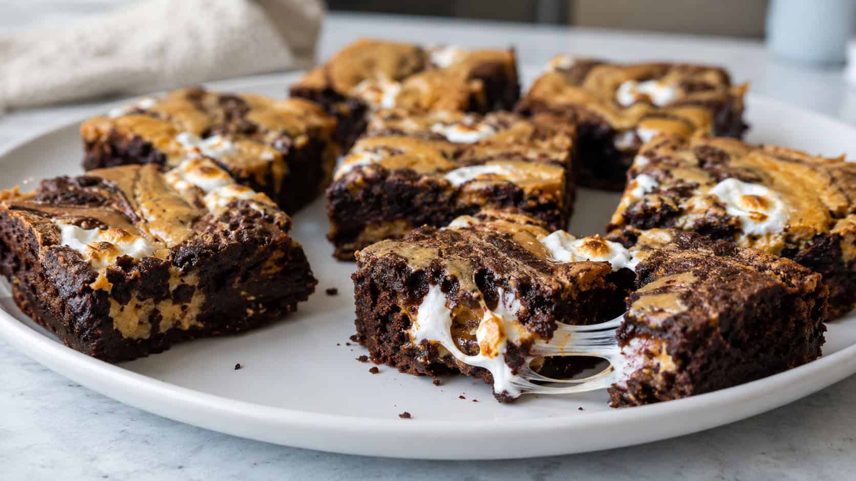 peanut butter marshmallow swirl brownies on a wide white plate in a horizontal shot, dark squares marbled with golden peanut butter and white toasted marshmallow ribbons, one bar pulled apart to show gooey swirls, crumbs on a soft-lit counter, natural light, shallow depth of field, landscape orienta