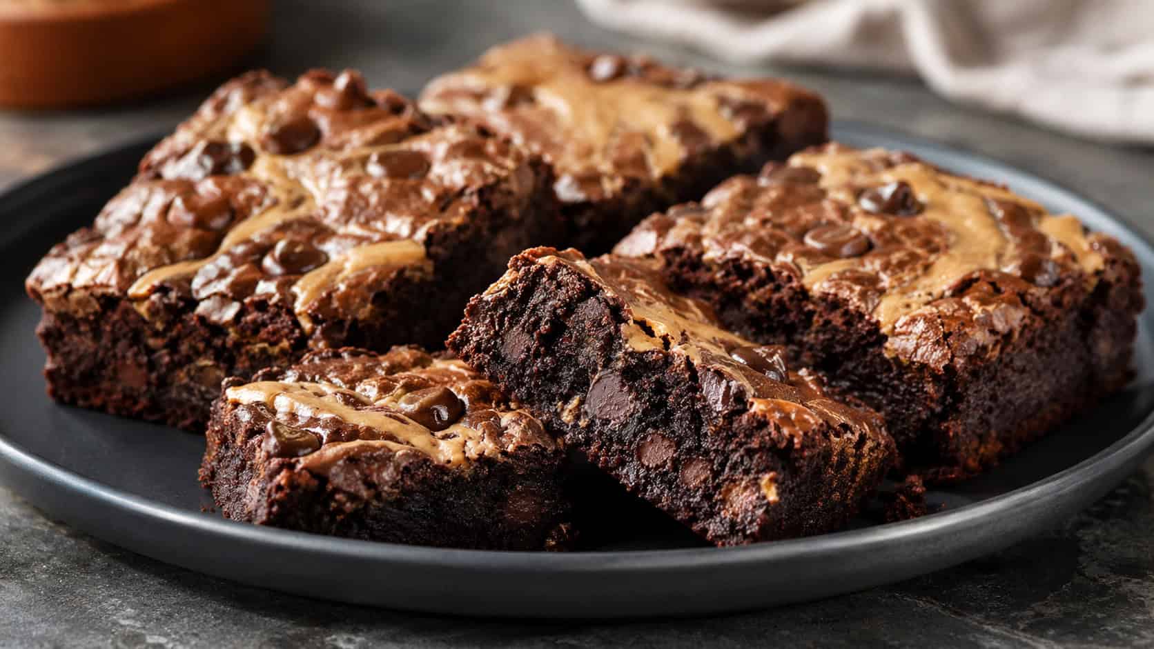 double chocolate peanut butter brownies on a wide dark plate in a horizontal shot, glossy squares with melty chocolate chips and golden peanut butter swirls, one bar broken to show a fudgy chip-studded crumb on a moody counter, warm natural light, shallow depth of field, landscape orientation