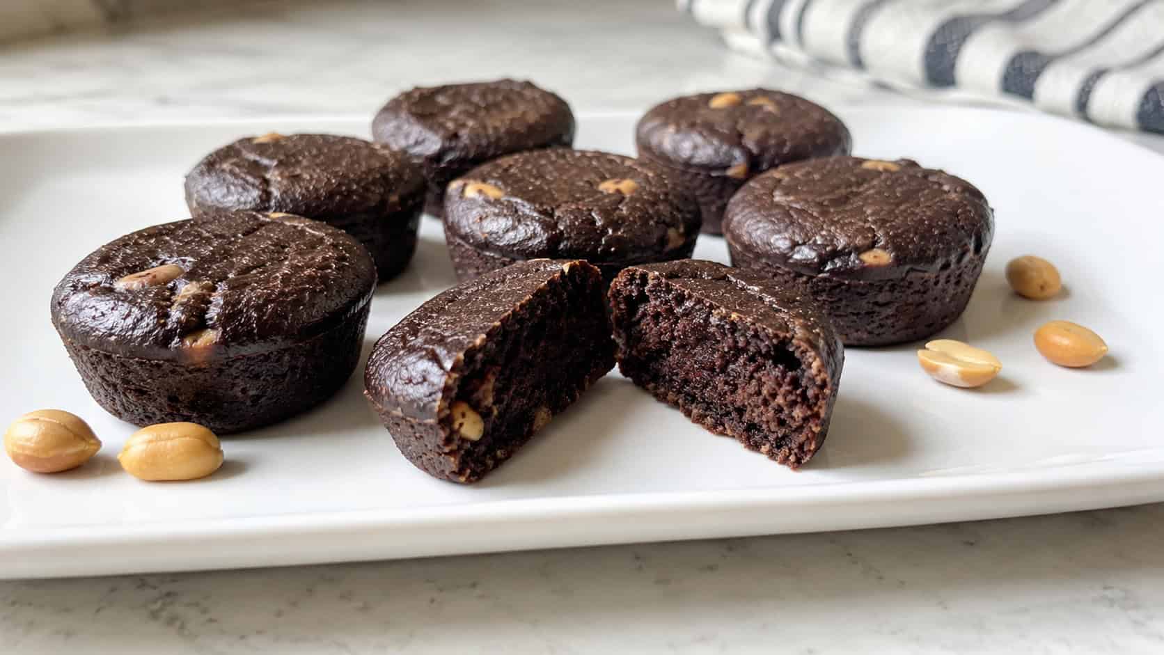 keto peanut butter brownie bites on a wide white plate in a horizontal shot, small dark fudgy rounds, one halved to show a moist crumb, a few peanuts scattered nearby on a clean bright counter, natural light, shallow depth of field, landscape orientation
