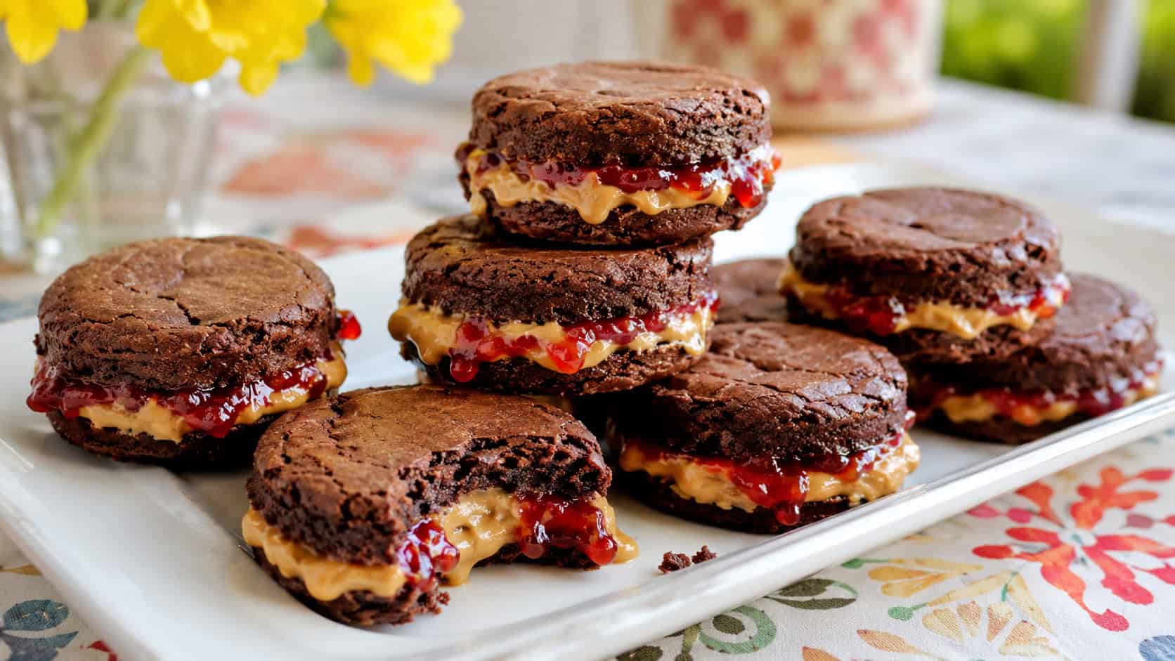 peanut butter and jelly brownie sliders on a wide platter in a horizontal shot, round brownie sandwiches oozing peanut butter and red jam from the middle, a few stacked and one bitten on a cheerful bright table, fresh natural light, shallow depth of field, landscape orientation