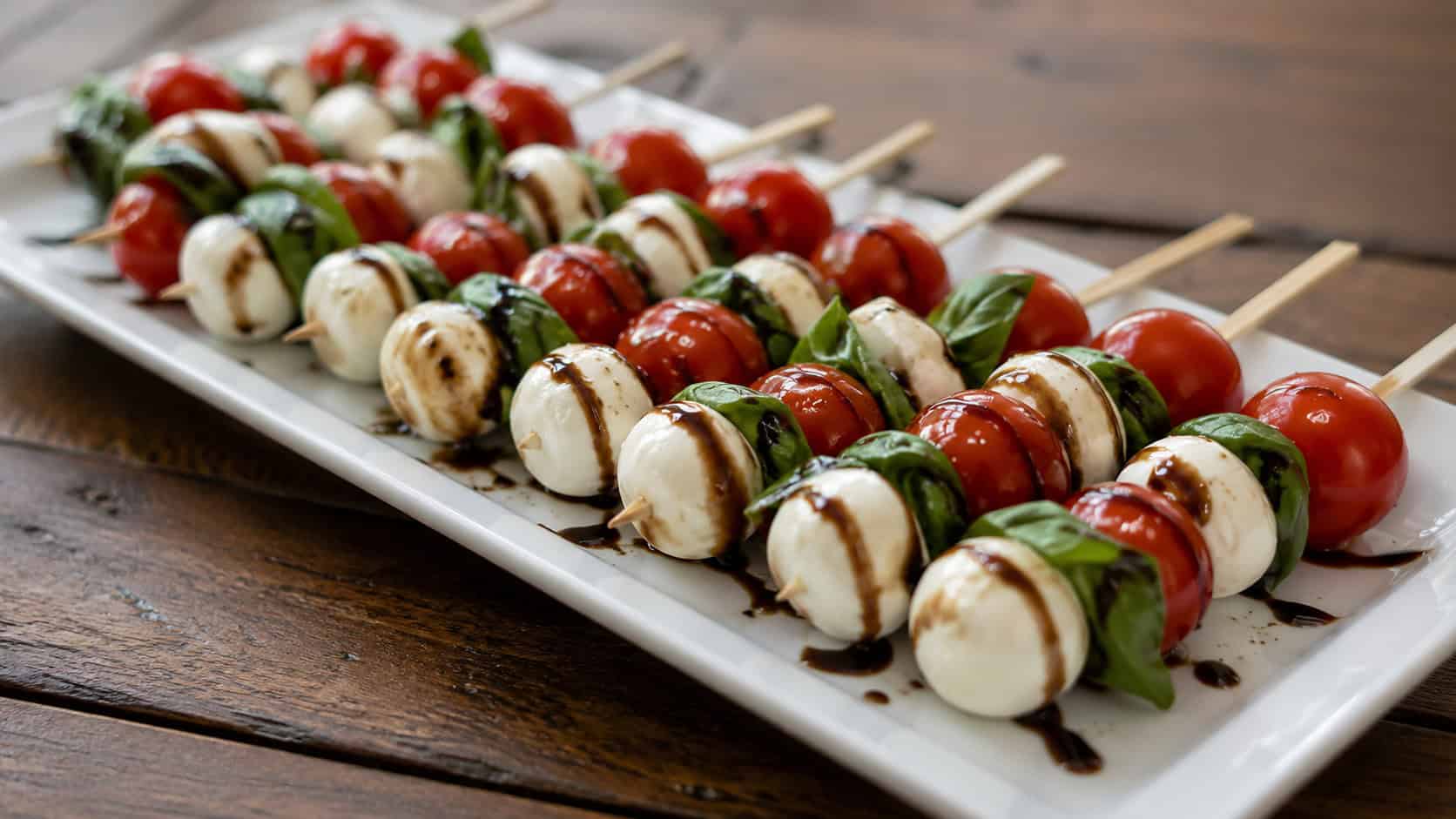rows of caprese skewers with tomato, basil, and mozzarella drizzled with balsamic glaze in a wide horizontal shot, on a white platter over a wooden table, natural light, shallow depth of field, landscape orientation