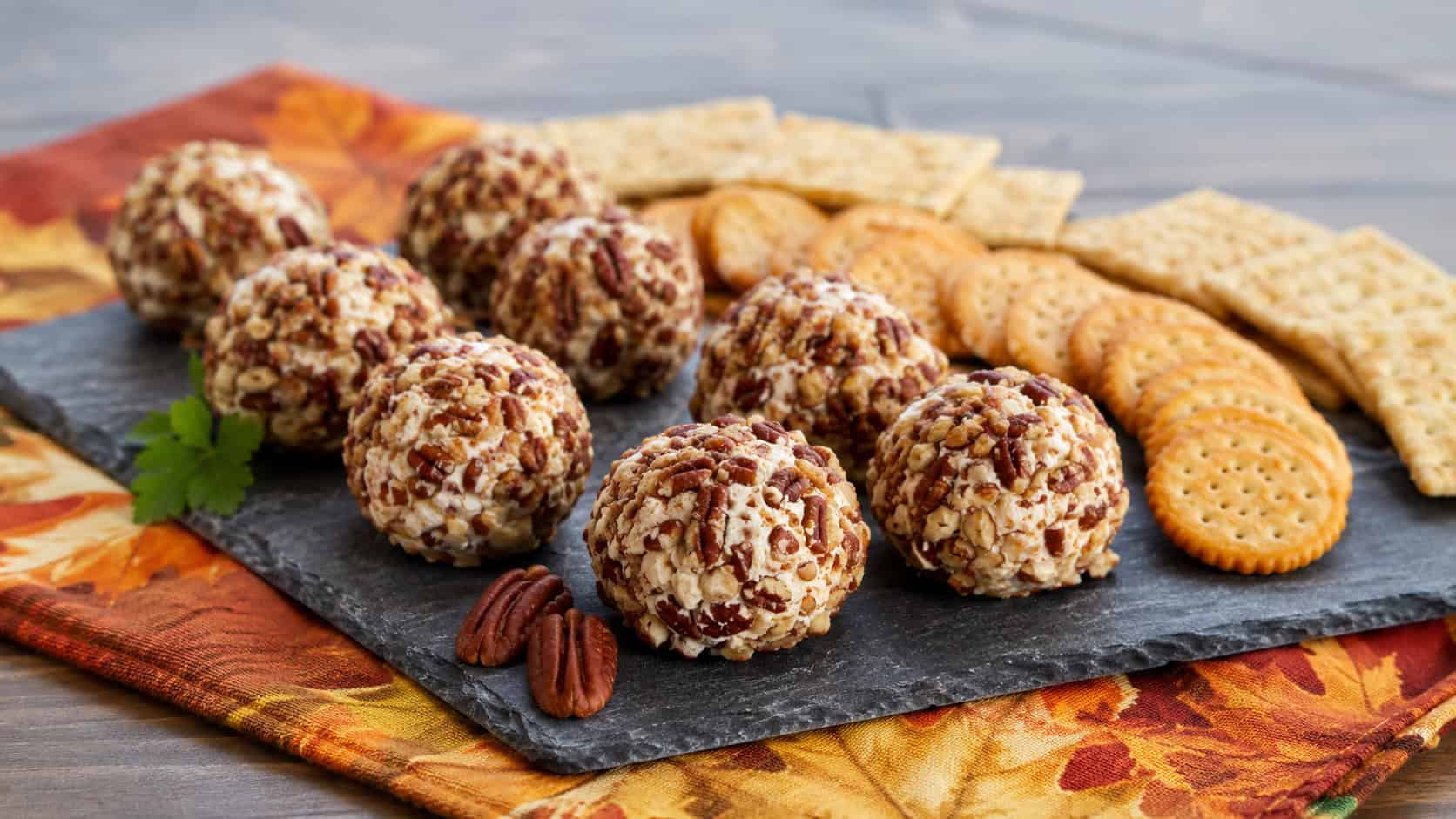 a platter of mini cheese balls coated in chopped pecans with crackers alongside in a wide horizontal shot, on a slate board over a fall napkin, natural light, shallow depth of field, landscape orientation