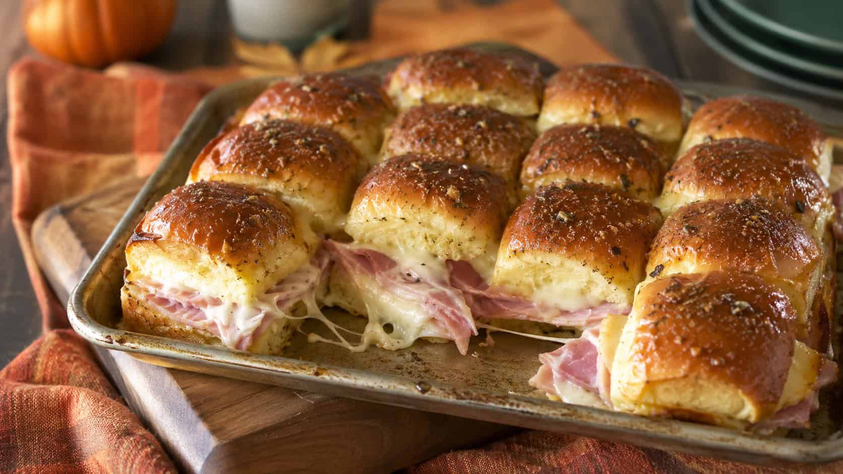 a tray of golden ham and cheese sliders glazed and pulled slightly apart in a wide horizontal shot, on a wooden board over a fall-toned cloth, natural light, shallow depth of field, landscape orientation