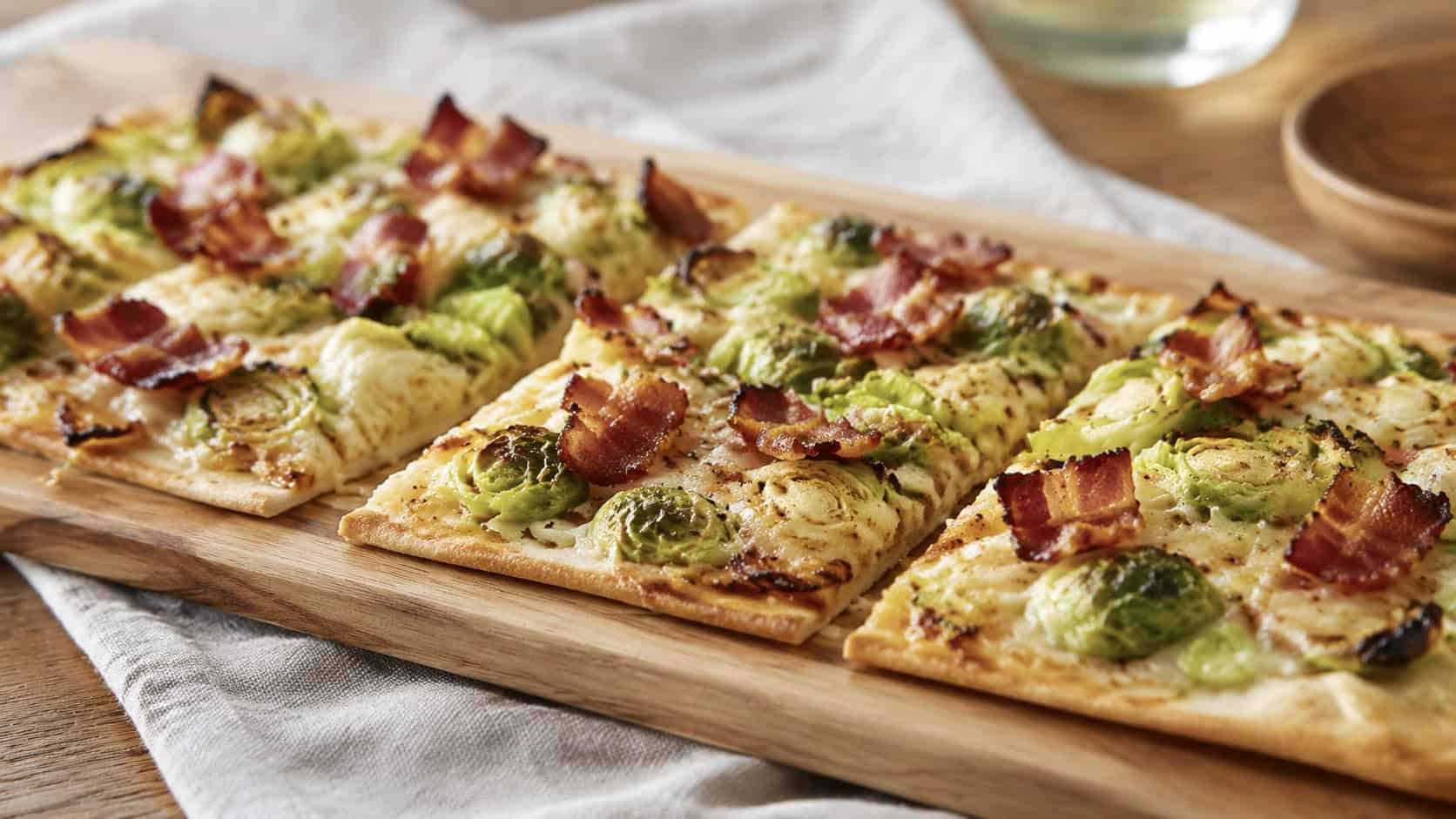 a crisp flatbread topped with shaved brussels sprouts, bacon, and melted cheese, sliced in a wide horizontal shot, on a wooden board over a linen napkin, natural light, shallow depth of field, landscape orientation