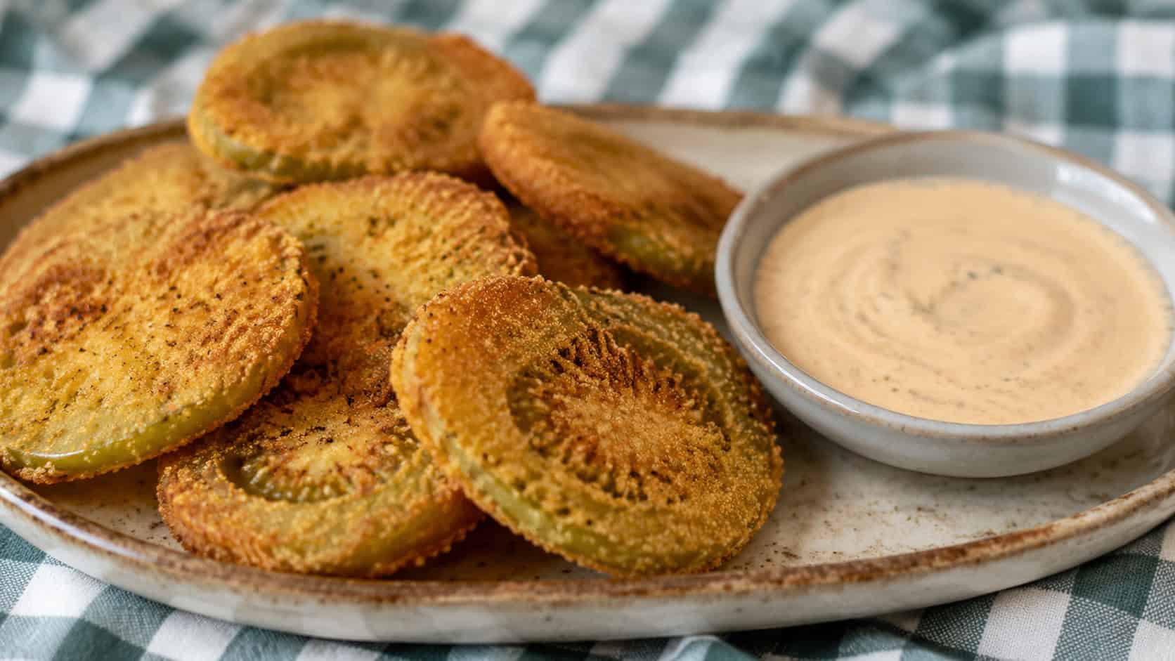 golden crispy fried green tomato slices stacked beside a creamy dip in a wide horizontal shot, on a rustic plate over a checkered cloth, natural light, shallow depth of field, landscape orientation