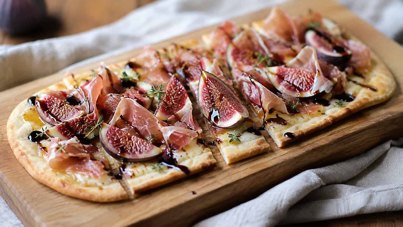 a crisp flatbread topped with fig slices, torn prosciutto, and balsamic drizzle, sliced in a wide horizontal shot, on a wooden board over a linen napkin, natural light, shallow depth of field, landscape orientation