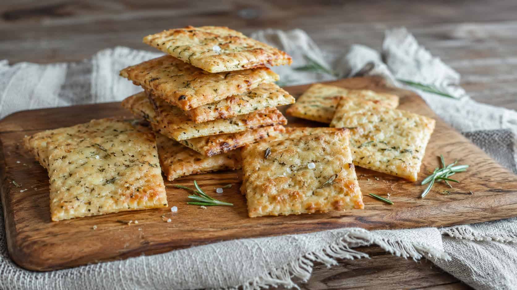 crisp homemade crackers flecked with rosemary stacked and scattered in a wide horizontal shot, on a wooden board over a rustic cloth, natural light, shallow depth of field, landscape orientation