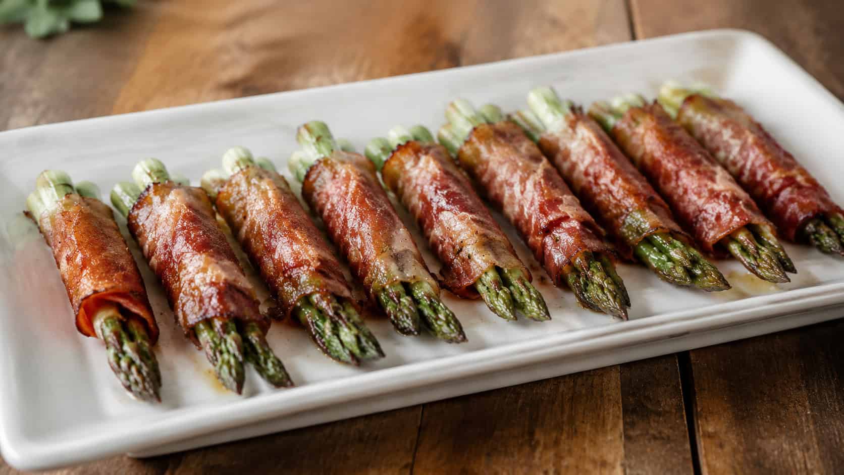roasted asparagus spears wrapped in crisp prosciutto arranged in a neat row in a wide horizontal shot, on a long white platter over a wooden table, natural light, shallow depth of field, landscape orientation