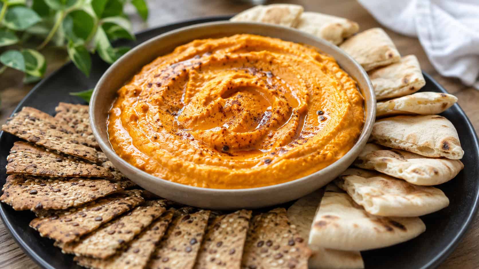 a bowl of creamy orange pumpkin hummus swirled and drizzled with oil beside pita in a wide horizontal shot, on a dark plate with crackers fanned around, natural light, shallow depth of field, landscape orientation