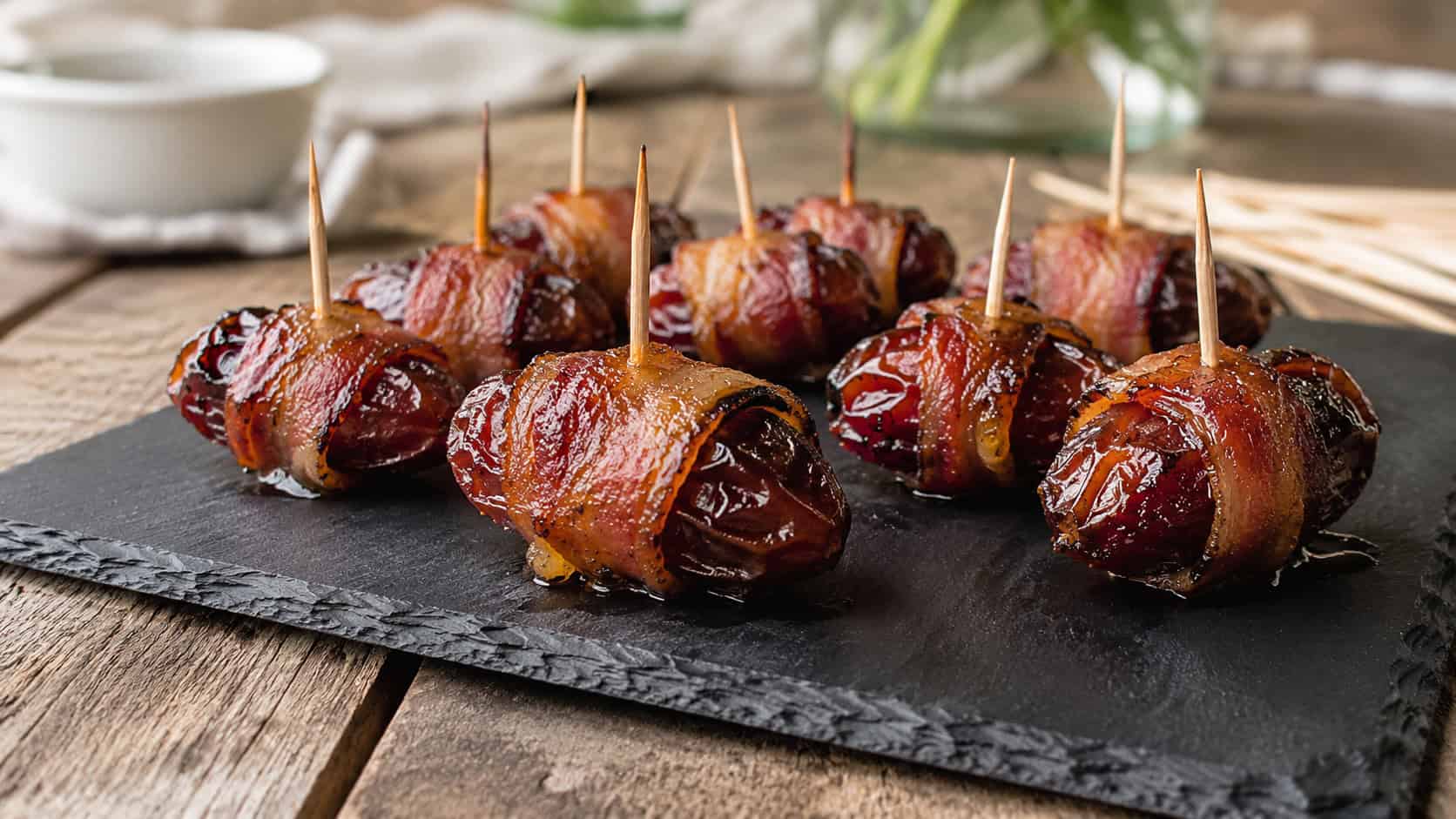 bacon-wrapped dates glazed with maple and skewered with toothpicks in a wide horizontal shot, on a slate board over a rustic table, natural light, shallow depth of field, landscape orientation