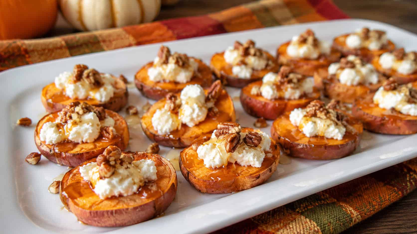 roasted sweet potato rounds topped with goat cheese, honey drizzle, and pecans in a wide horizontal shot, on a white platter over a fall-toned napkin, natural light, shallow depth of field, landscape orientation