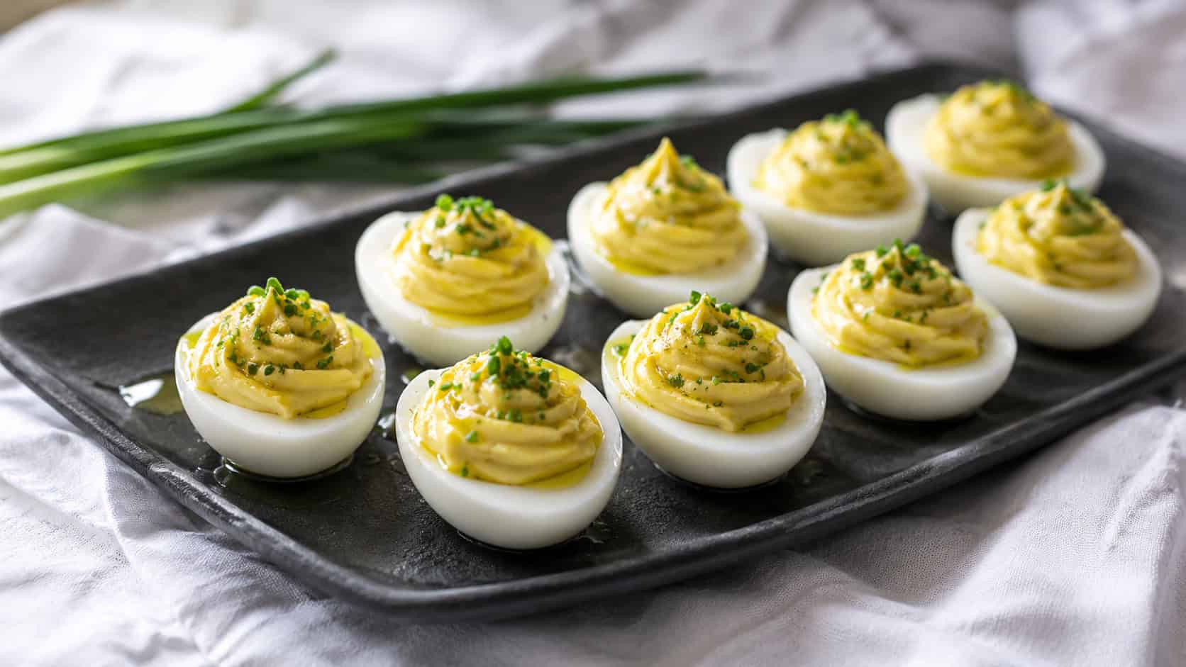 deviled eggs piped smooth and topped with chives and a glint of oil in a wide horizontal shot, on a dark platter over a white cloth, natural light, shallow depth of field, landscape orientation