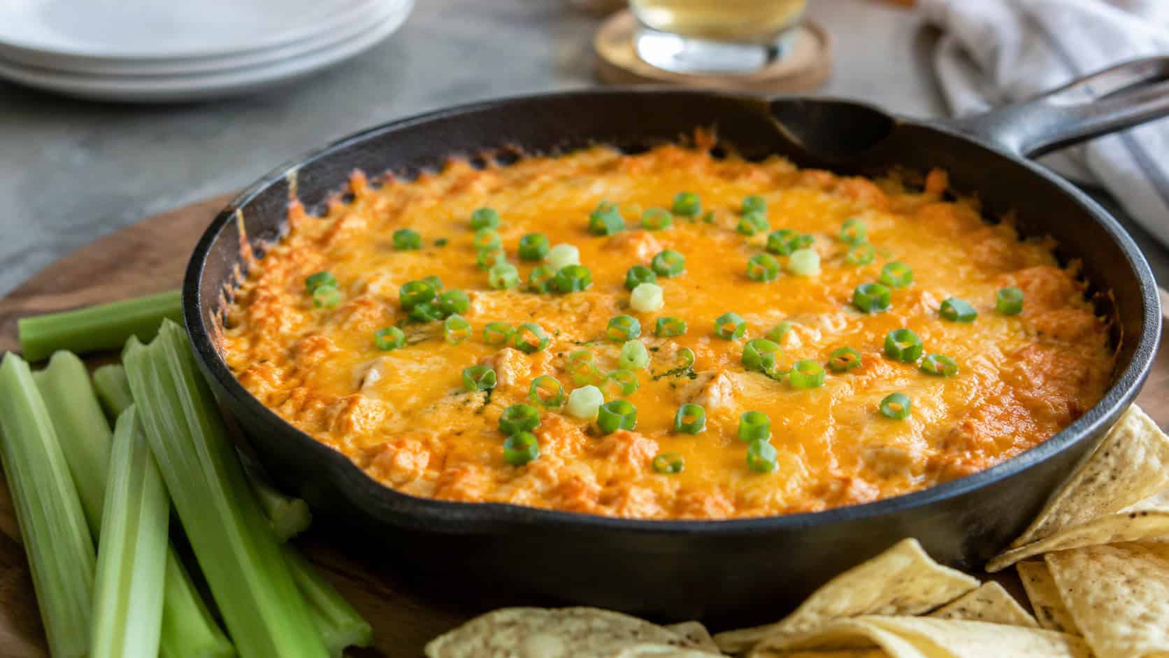 a bubbling buffalo chicken dip topped with melted cheddar and green onions in a wide horizontal shot, served in a cast iron skillet with celery sticks and chips, natural light, shallow depth of field, landscape orientation