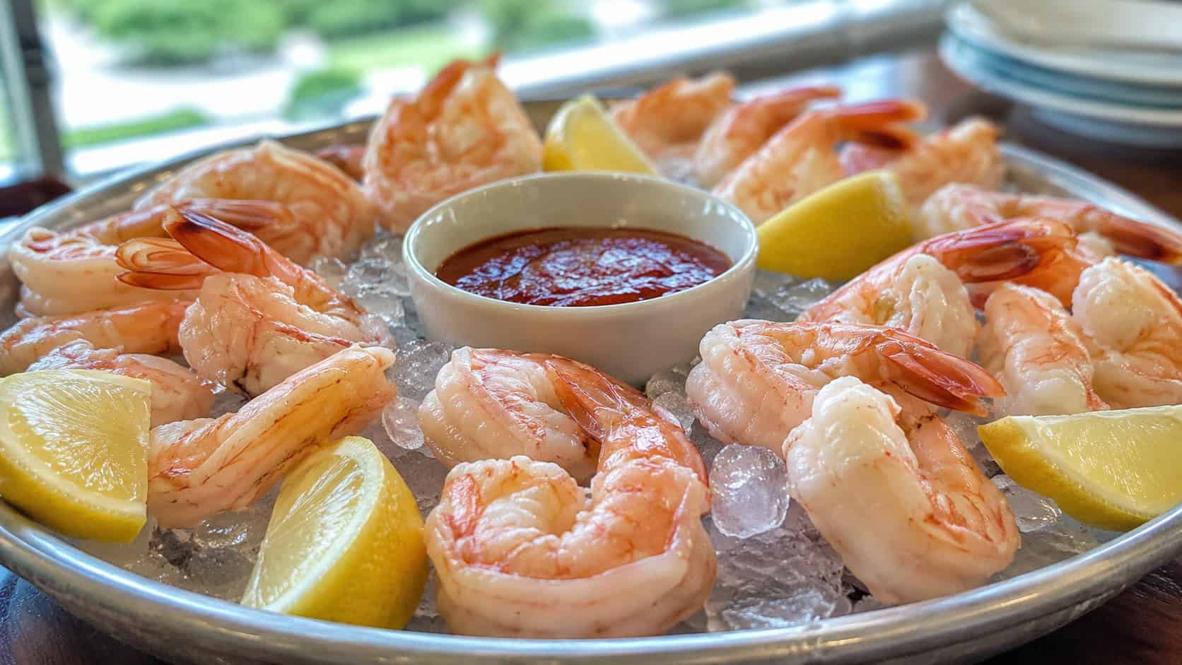 chilled pink shrimp arranged around a bowl of red cocktail sauce in a wide horizontal shot, served on a bed of ice with lemon wedges, natural light, shallow depth of field, landscape orientation