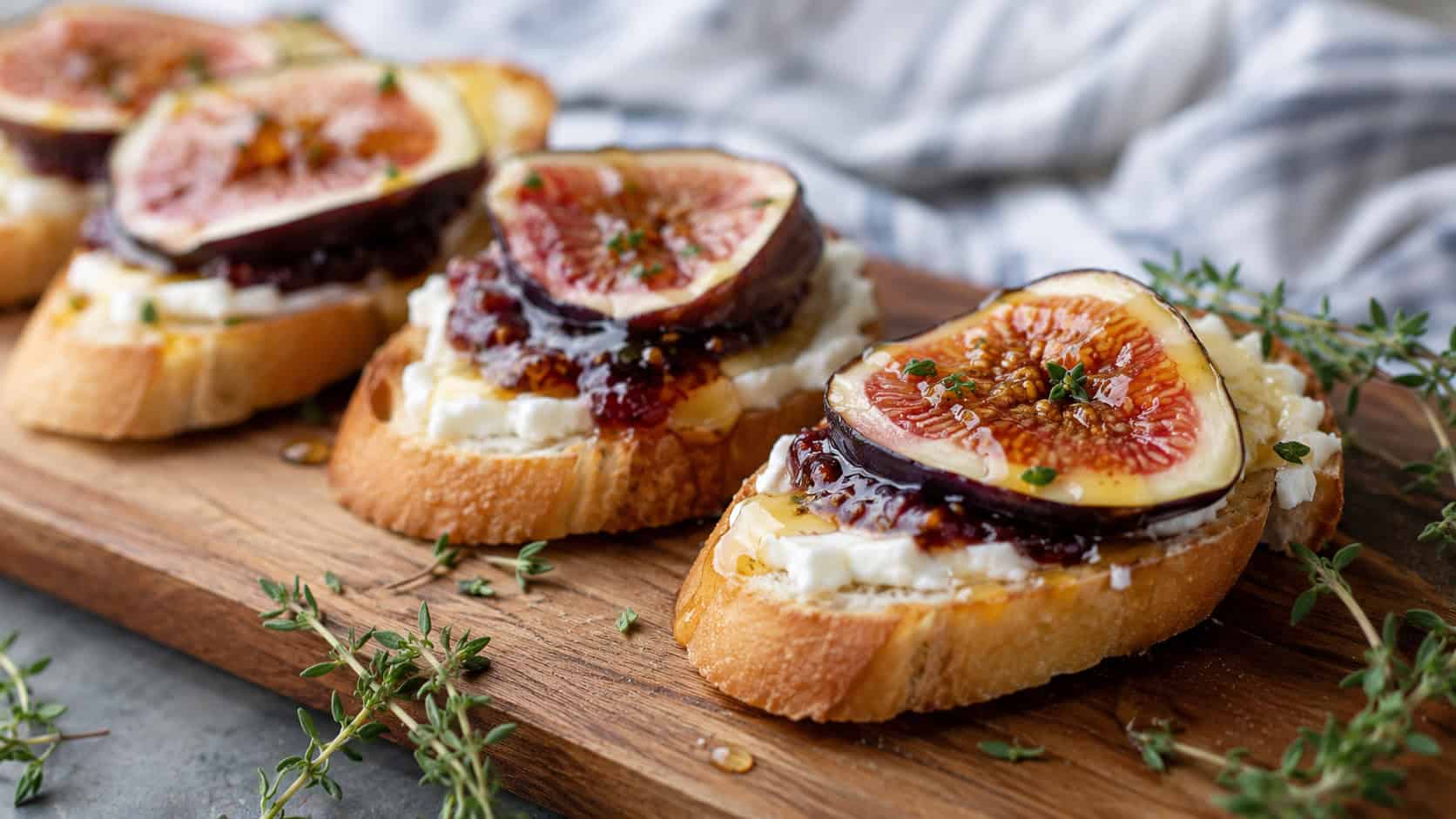 fig and goat cheese crostini with creamy cheese, jam, and honey drizzle in a wide horizontal shot, lined up on a wooden serving board with thyme sprigs, natural light, shallow depth of field, landscape orientation
