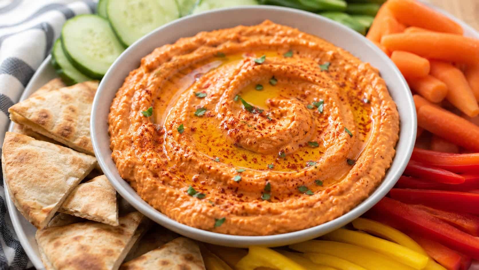 a bowl of creamy roasted red pepper hummus swirled with olive oil and paprika in a wide horizontal shot, surrounded by pita wedges and fresh vegetables, natural light, shallow depth of field, landscape orientation
