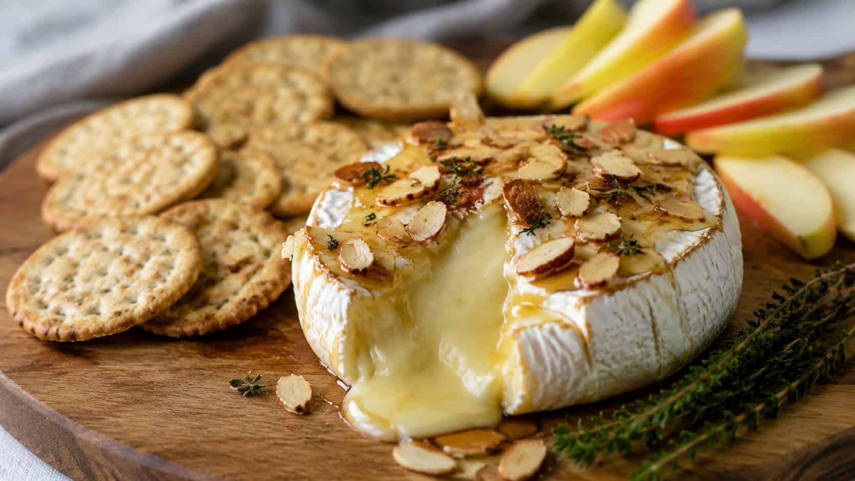 a warm wheel of baked brie oozing in the center, topped with honey and toasted almonds in a wide horizontal shot, served with crackers and apple slices on a board, natural light, shallow depth of field, landscape orientation