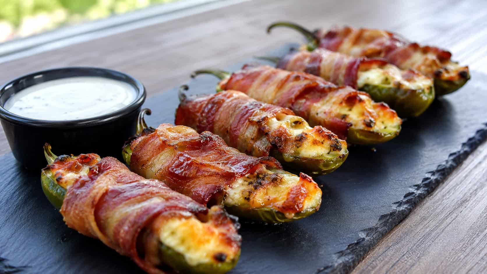 a row of bacon-wrapped jalapeño poppers with crisp bacon and bubbling cheese filling in a wide horizontal shot, served on a slate board with a small dish of ranch, natural light, shallow depth of field, landscape orientation