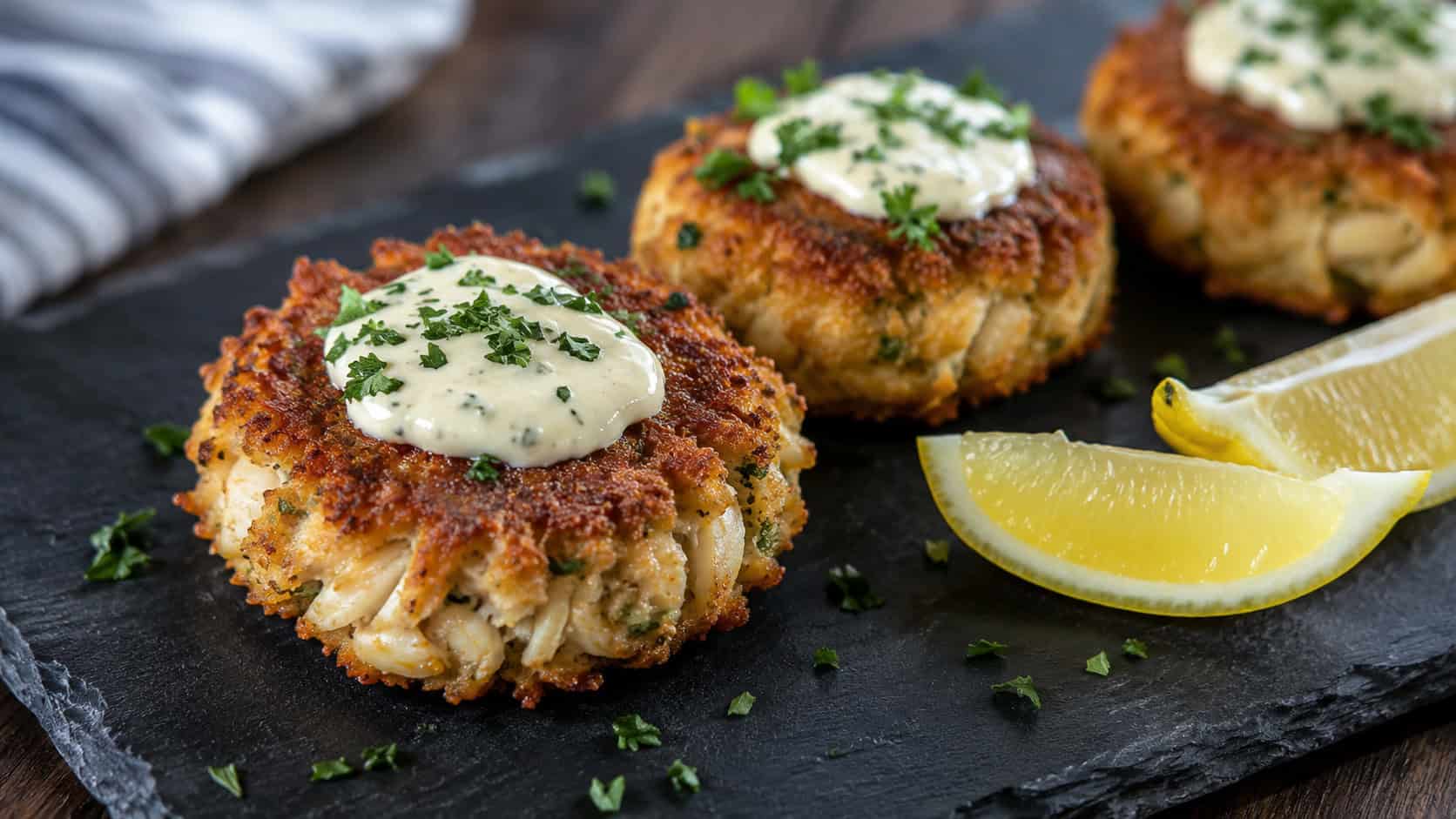 golden crispy crab cakes topped with creamy aioli and parsley in a wide horizontal shot, plated with lemon wedges on a dark slate board, natural light, shallow depth of field, landscape orientation