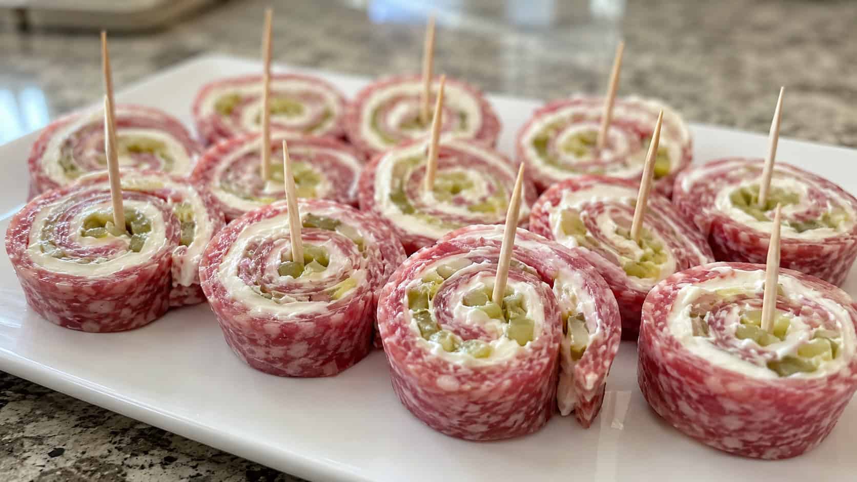 salami and cream cheese pinwheels showing spirals of meat, cream cheese, and pickle in a wide horizontal shot, arranged on a white platter with toothpicks, natural light, shallow depth of field, landscape orientation