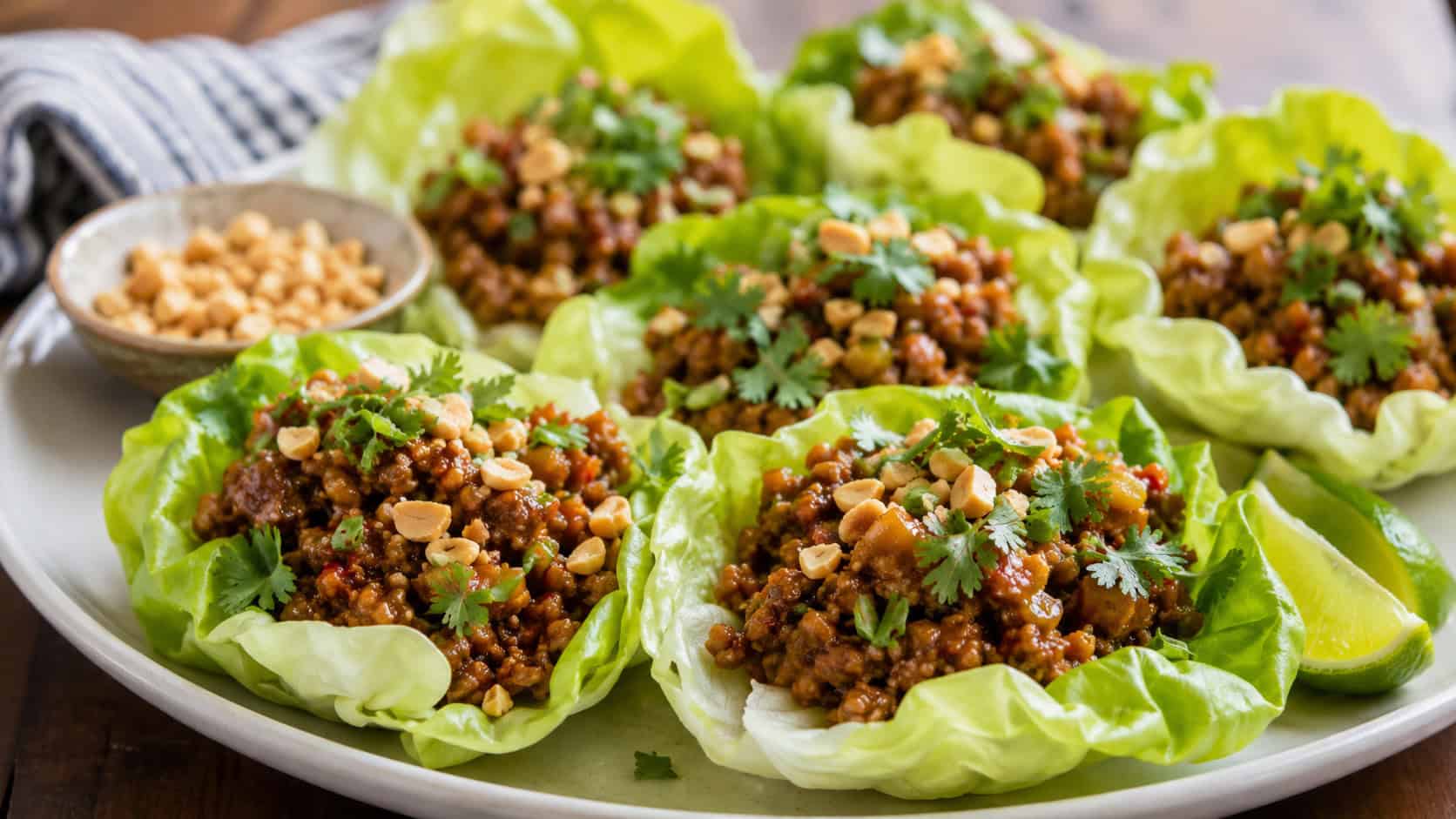 Thai chicken lettuce cups filled with saucy ground chicken, peanuts, and cilantro in a wide horizontal shot, arranged on a platter with lime wedges, natural light, shallow depth of field, landscape orientation