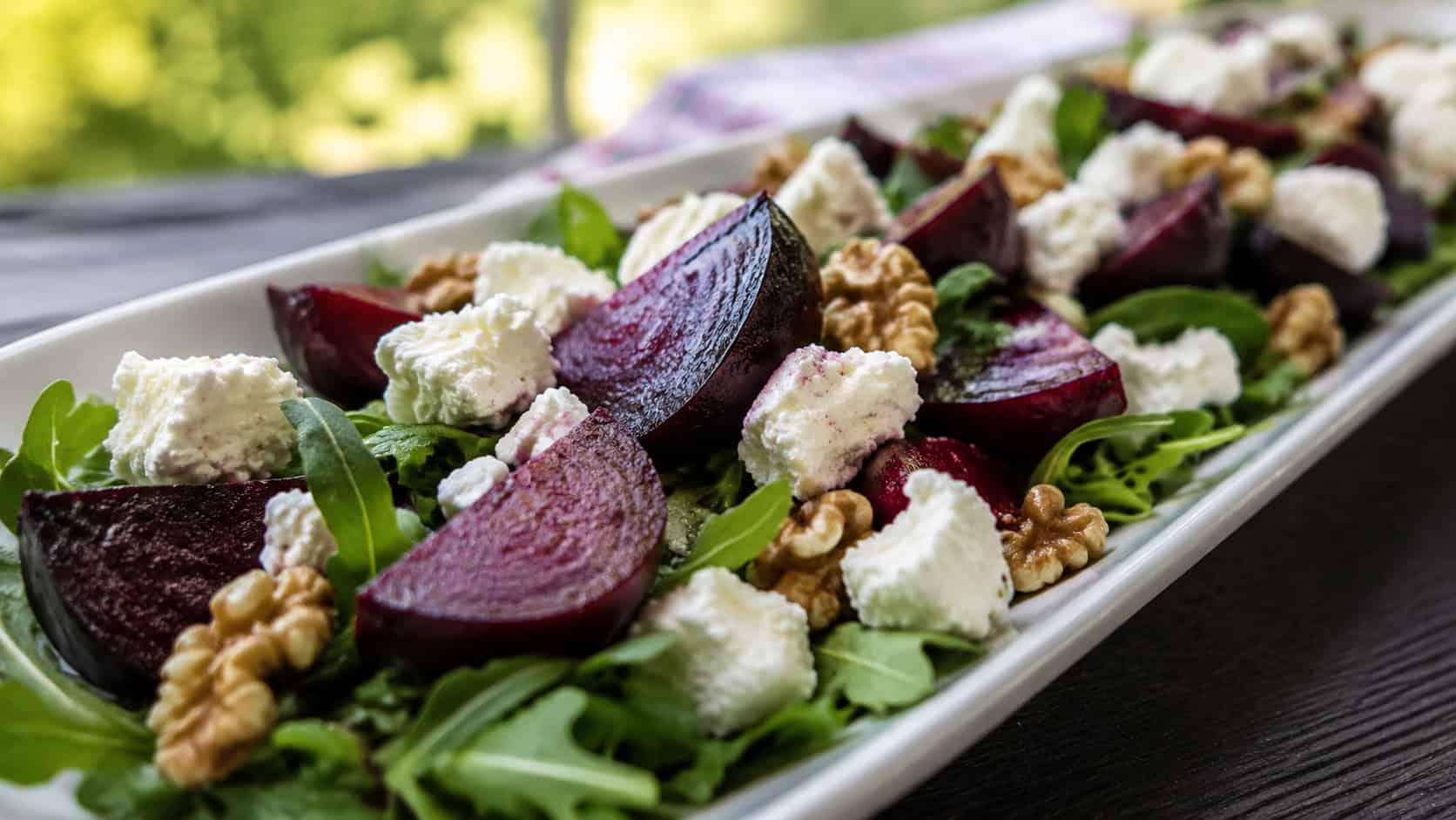 a beet and goat cheese salad with ruby beet wedges, creamy cheese, and walnuts over greens in a wide horizontal shot, served on a long platter, natural light, shallow depth of field, landscape orientation