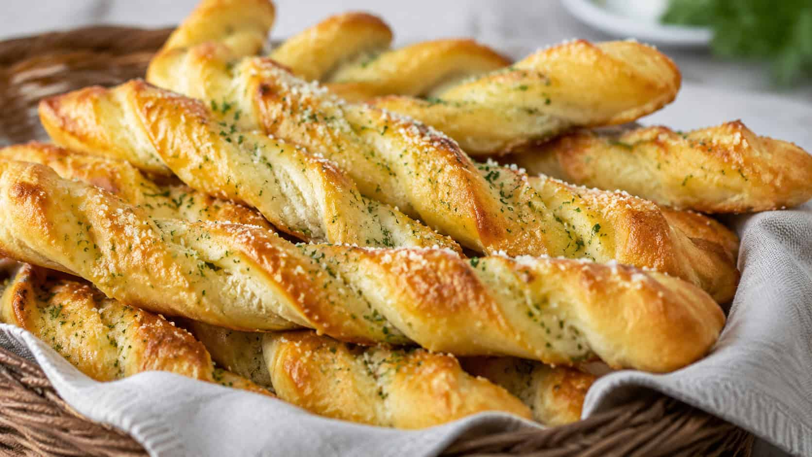 golden twisted garlic breadsticks brushed with butter and dusted with Parmesan in a wide horizontal shot, arranged in a basket lined with a cloth napkin, natural light, shallow depth of field, landscape orientation