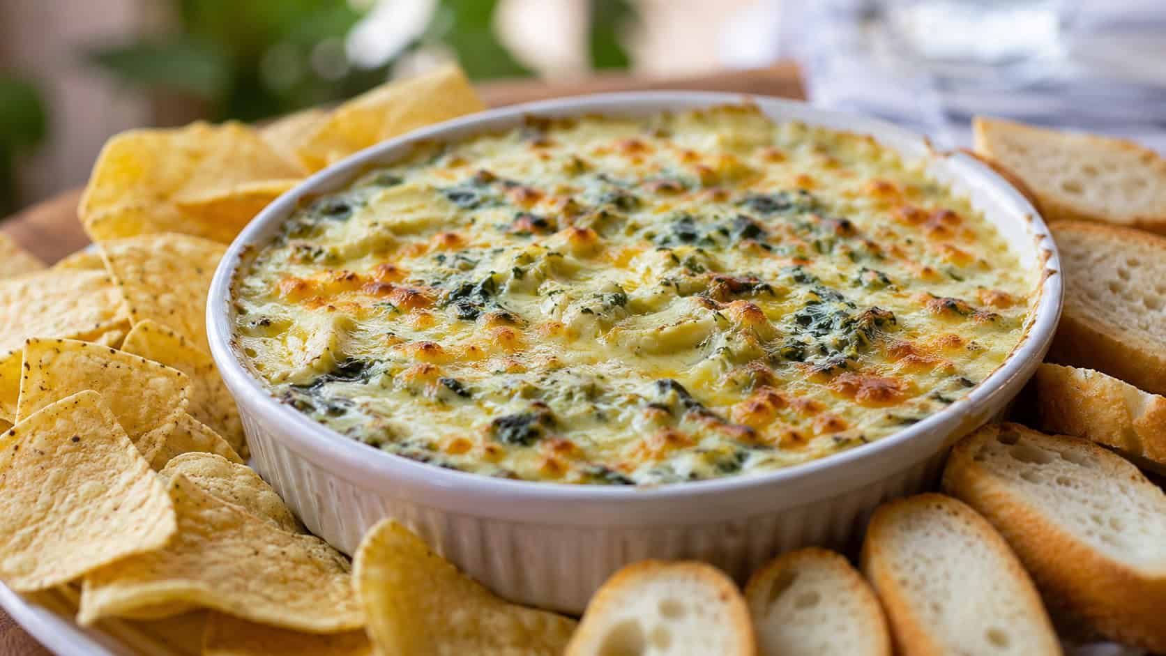 a bubbling golden spinach and artichoke dip in a wide horizontal shot, served in a round baking dish surrounded by tortilla chips and sliced bread, natural light, shallow depth of field, landscape orientation