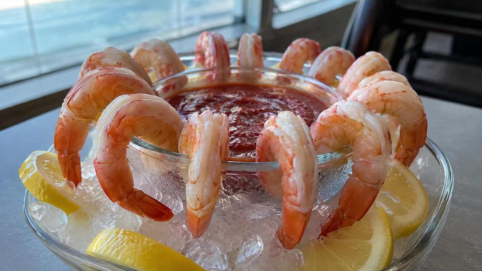 chilled shrimp cocktail in a wide horizontal shot, shrimp hung around the rim of a glass bowl of red sauce with lemon wedges on a bed of ice, natural light, shallow depth of field, landscape orientation