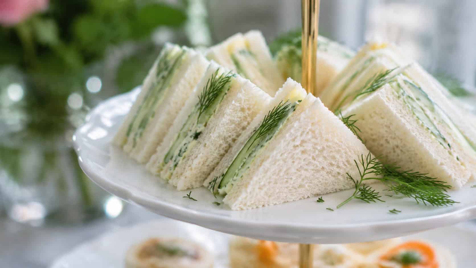 delicate cucumber tea sandwiches in a wide horizontal shot, cut into triangles and stacked on a tiered white stand with fresh dill, natural light, shallow depth of field, landscape orientation