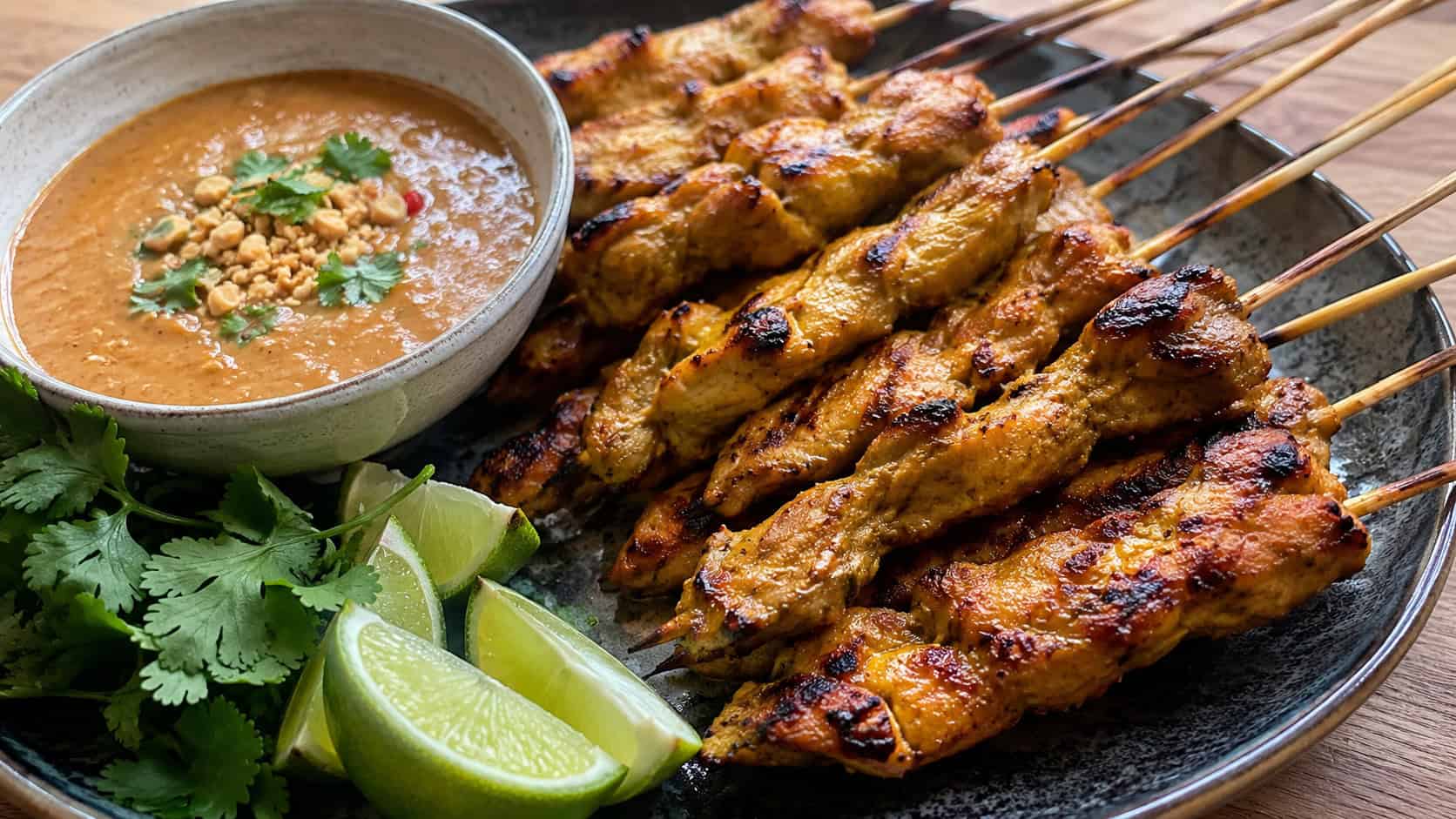 grilled chicken satay skewers in a wide horizontal shot, fanned on a platter beside a bowl of peanut sauce with lime wedges and cilantro, natural light, shallow depth of field, landscape orientation