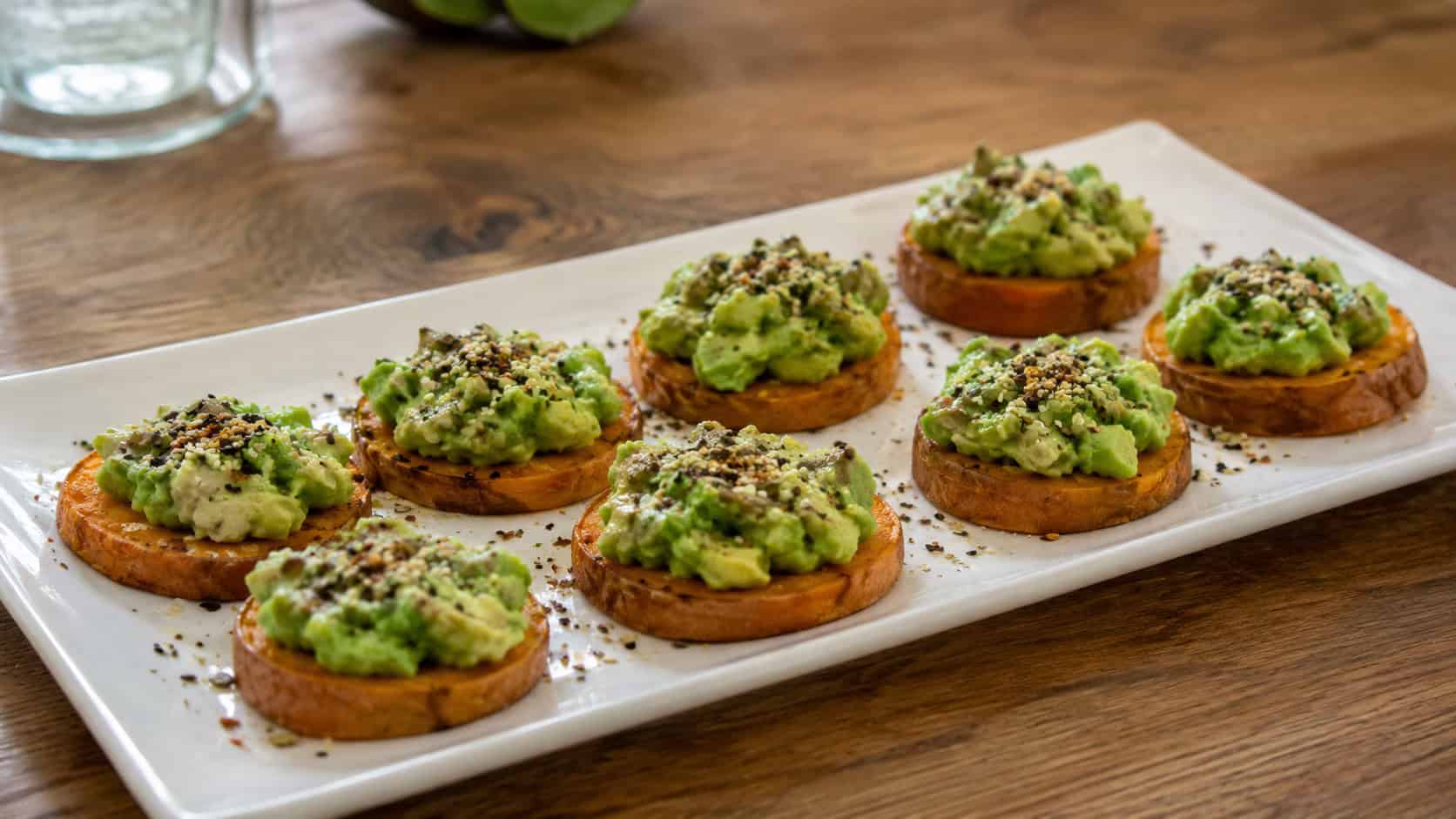 roasted sweet potato rounds topped with green avocado in a wide horizontal shot, arranged on a white platter sprinkled with seeds on a wooden table, natural light, shallow depth of field, landscape orientation