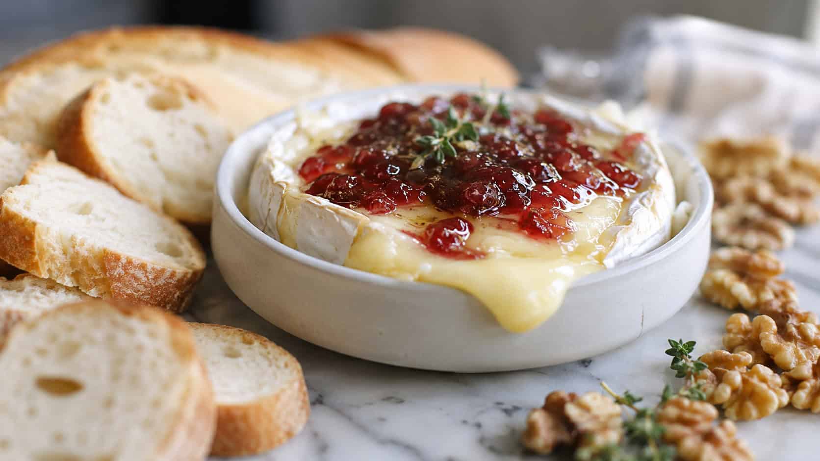 oozy baked brie topped with red jam in a wide horizontal shot, in a small dish surrounded by baguette slices and walnuts on a marble board, natural light, shallow depth of field, landscape orientation