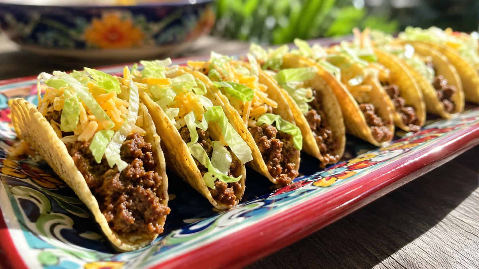 loaded mini tacos in a wide horizontal shot, lined up in a row filled with beef, cheese, and lettuce on a colorful platter, natural light, shallow depth of field, landscape orientation