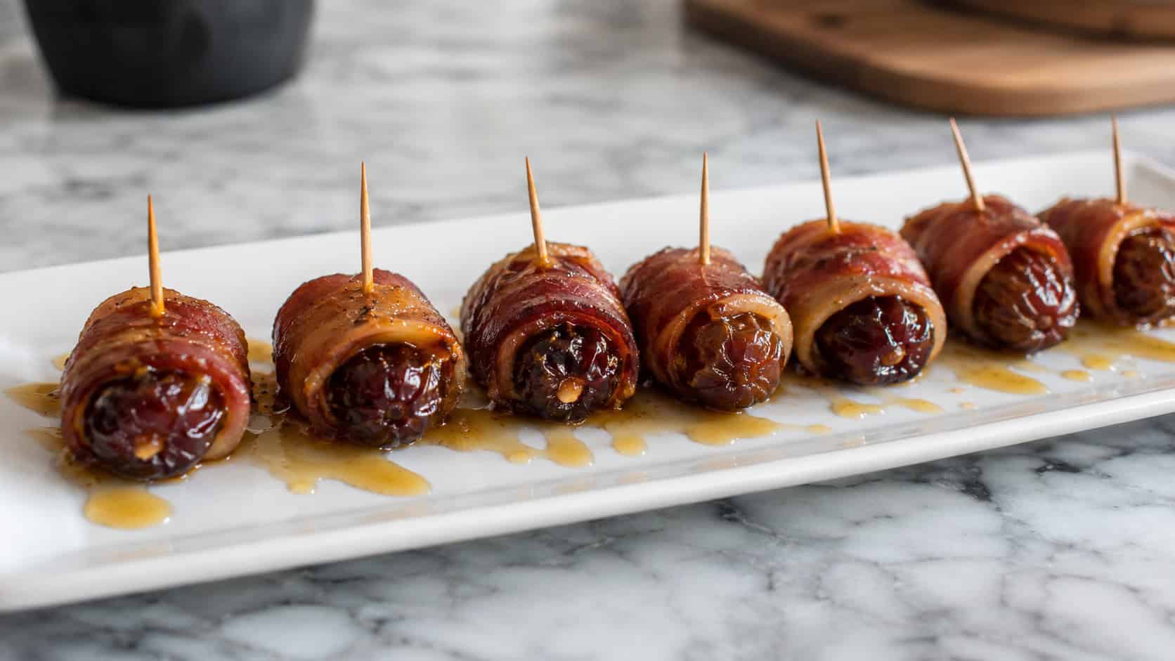 glossy bacon-wrapped dates in a wide horizontal shot, lined up on a white serving plate with toothpicks and a honey drizzle on a marble counter, natural light, shallow depth of field, landscape orientation