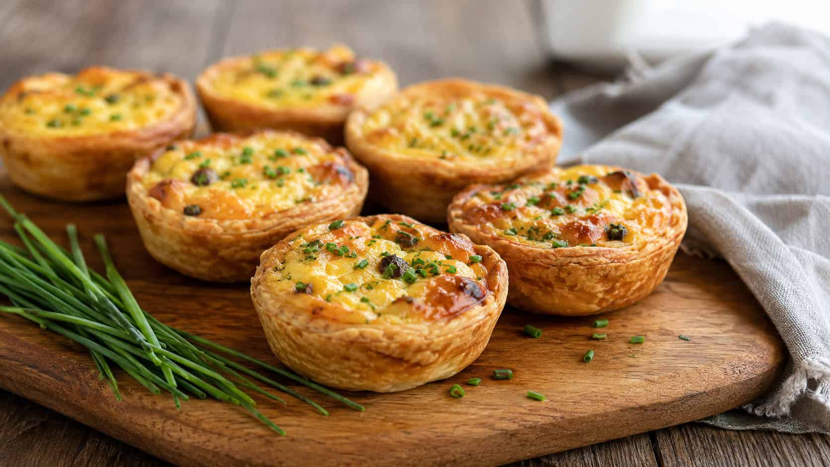 golden mini quiches in a wide horizontal shot, arranged on a wooden board with fresh chives and a linen napkin, natural light, shallow depth of field, landscape orientation