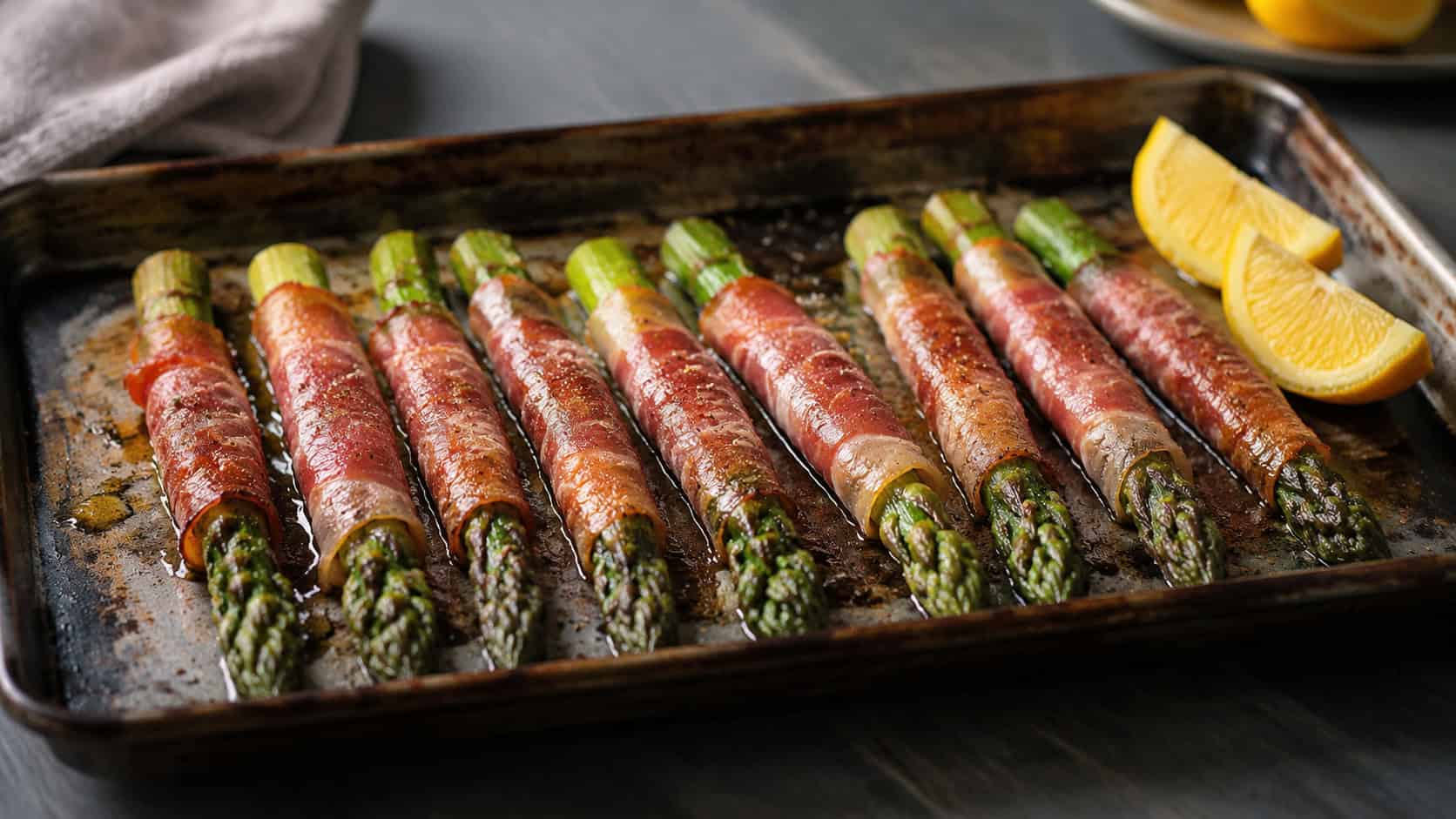 prosciutto-wrapped asparagus spears in a wide horizontal shot, lined up on a rustic baking tray with lemon wedges on a dark table, natural light, shallow depth of field, landscape orientation