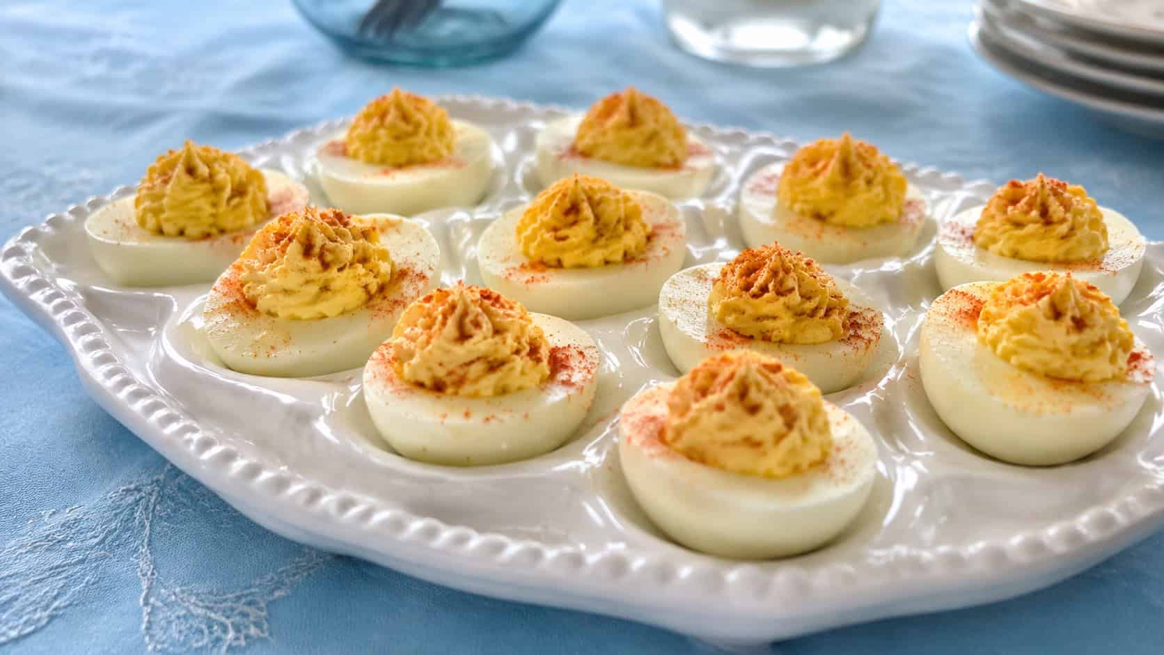 a tray of deviled eggs in a wide horizontal shot, dusted with paprika and arranged on a white deviled egg platter on a pale blue tablecloth, natural light, shallow depth of field, landscape orientation