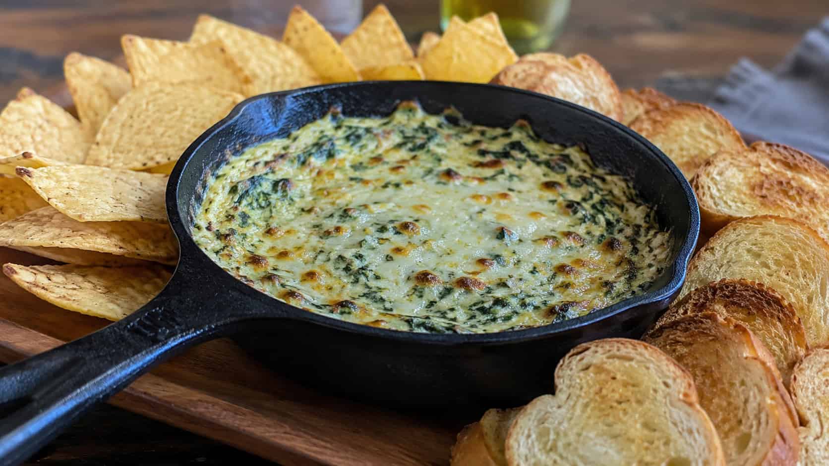 bubbling spinach and artichoke dip in a wide horizontal shot, in a cast iron skillet surrounded by toasted bread and tortilla chips on a wooden table, natural light, shallow depth of field, landscape orientation