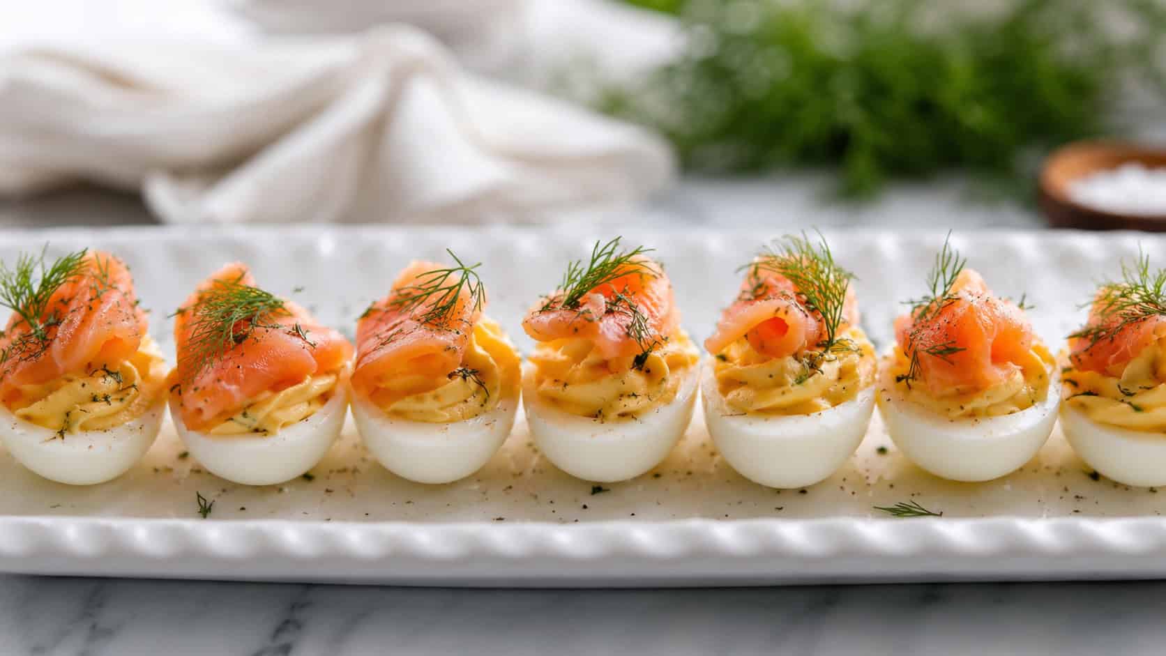 deviled eggs topped with smoked salmon and dill, arranged in a wide horizontal row on a white platter, elegant and fresh, soft natural light, shallow depth of field, landscape orientation