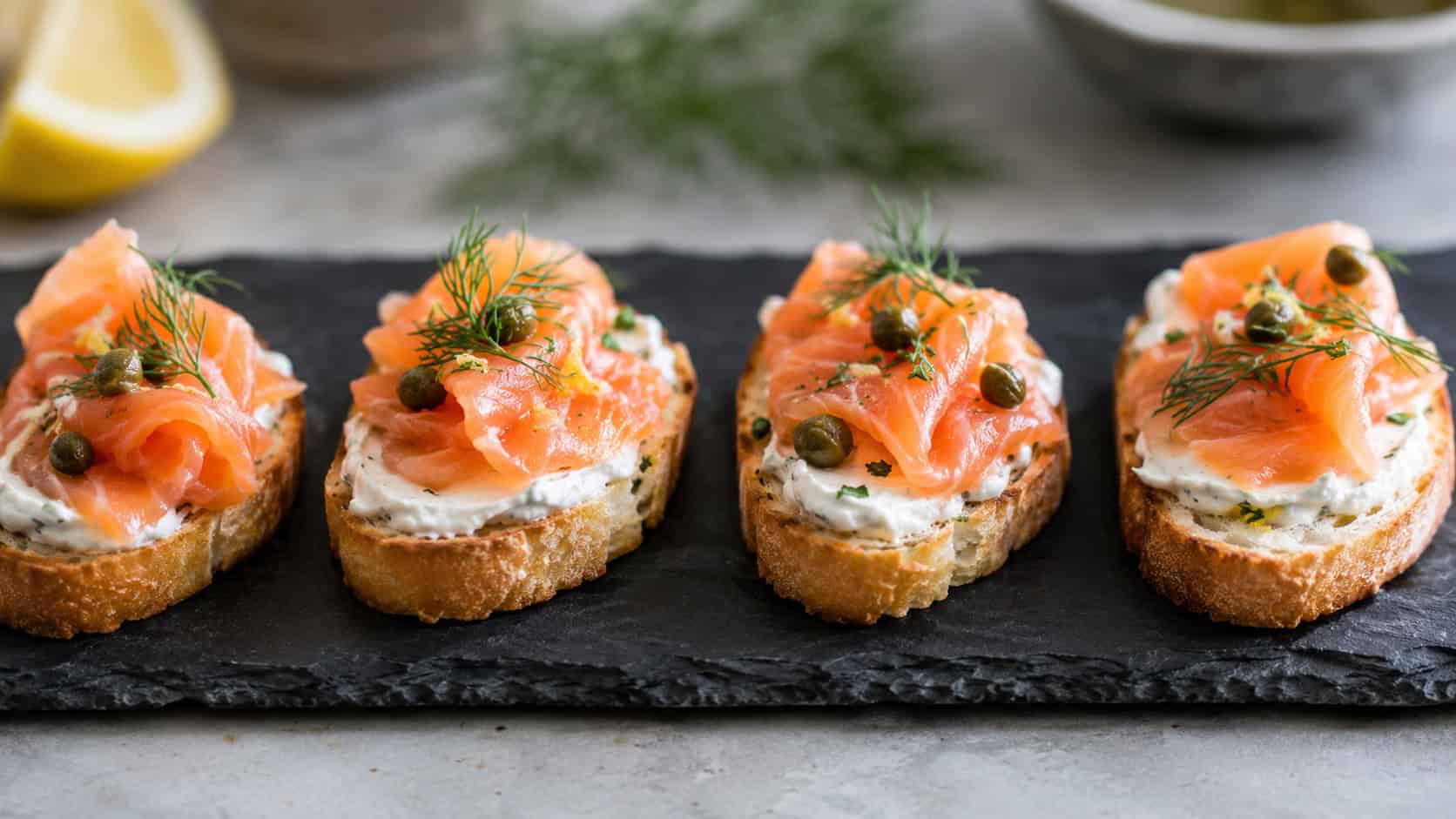 smoked salmon crostini with creamy spread, pink salmon, capers, and dill, arranged in a wide horizontal row on a slate board, elegant and fresh, soft natural light, shallow depth of field, landscape orientation