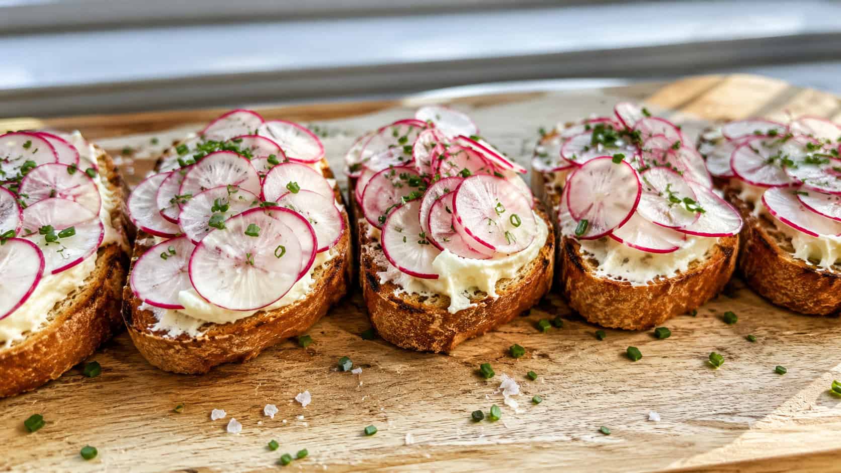 radish toasts with whipped butter and thin pink-edged radish slices sprinkled with chives and flaky salt, arranged in a wide horizontal row on a wooden board, bright natural light, shallow depth of field, landscape orientation