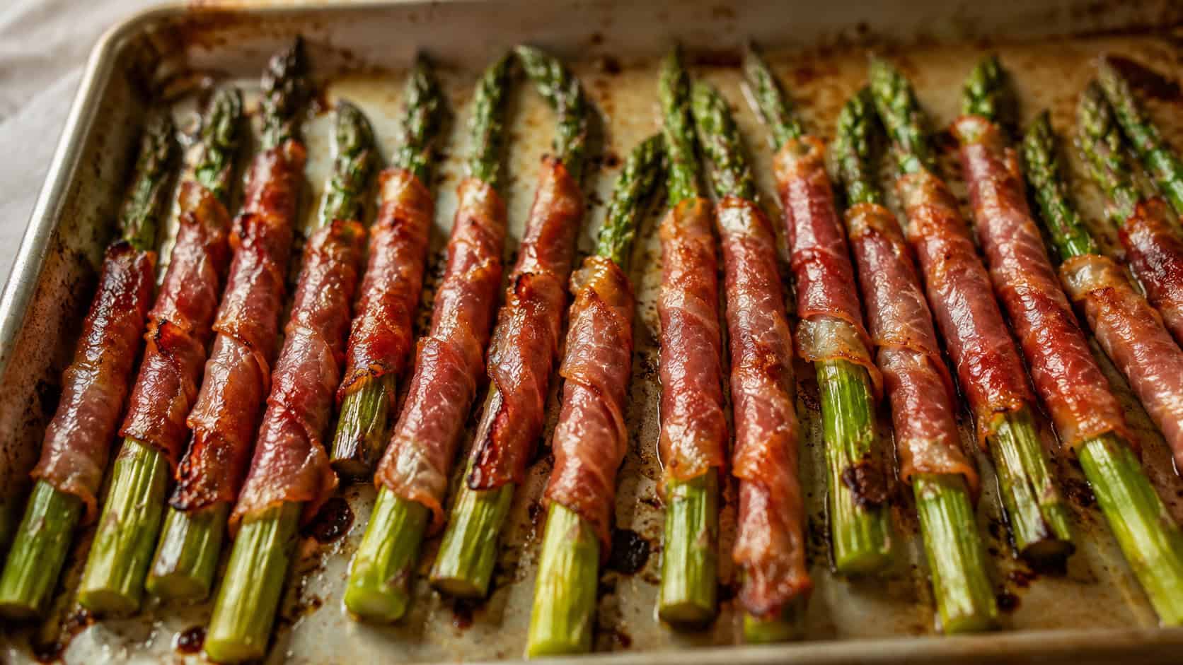 prosciutto-wrapped asparagus spears with crisp golden edges, arranged in a wide horizontal row on a sheet pan, glistening with olive oil, warm natural light, shallow depth of field, landscape orientation