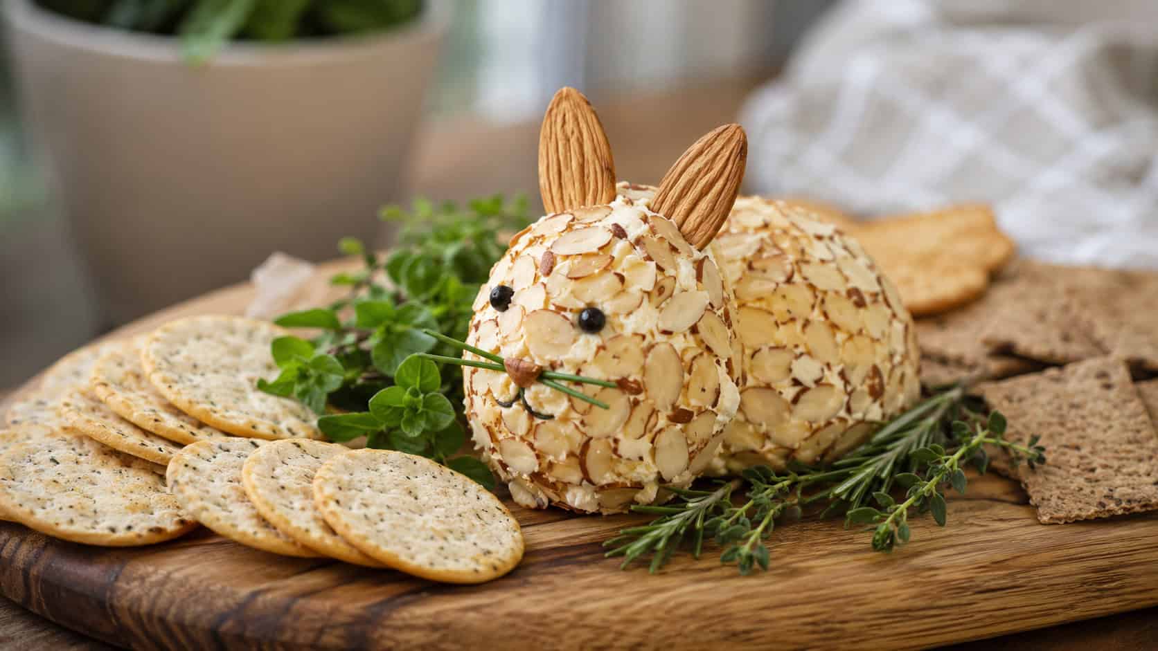 a bunny-shaped cheese ball coated in slivered almonds with almond ears, on a wide horizontal wooden board surrounded by crackers and fresh herbs, soft natural light, shallow depth of field, landscape orientation