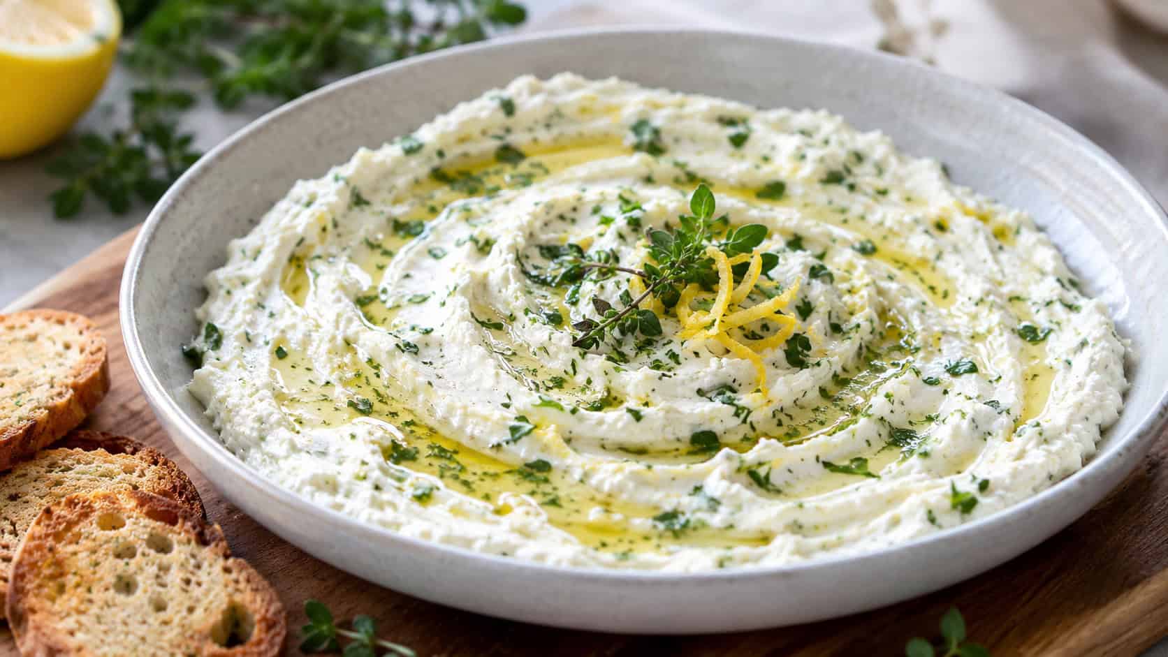 a swirled lemon herb goat cheese spread drizzled with olive oil and topped with fresh herbs, in a wide horizontal shallow bowl on a wooden board with crostini, bright natural light, shallow depth of field, landscape orientation