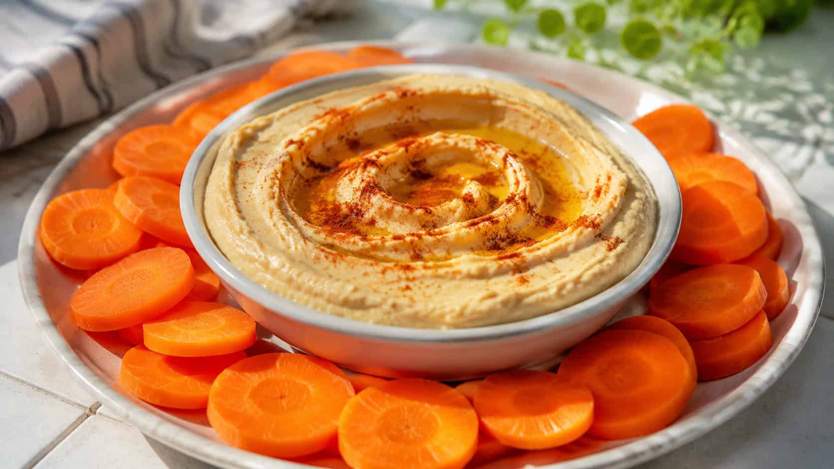 a swirled bowl of creamy hummus drizzled with olive oil and paprika, surrounded by crisp orange carrot coins, in a wide horizontal shot on a bright table, natural daylight, shallow depth of field, landscape orientation