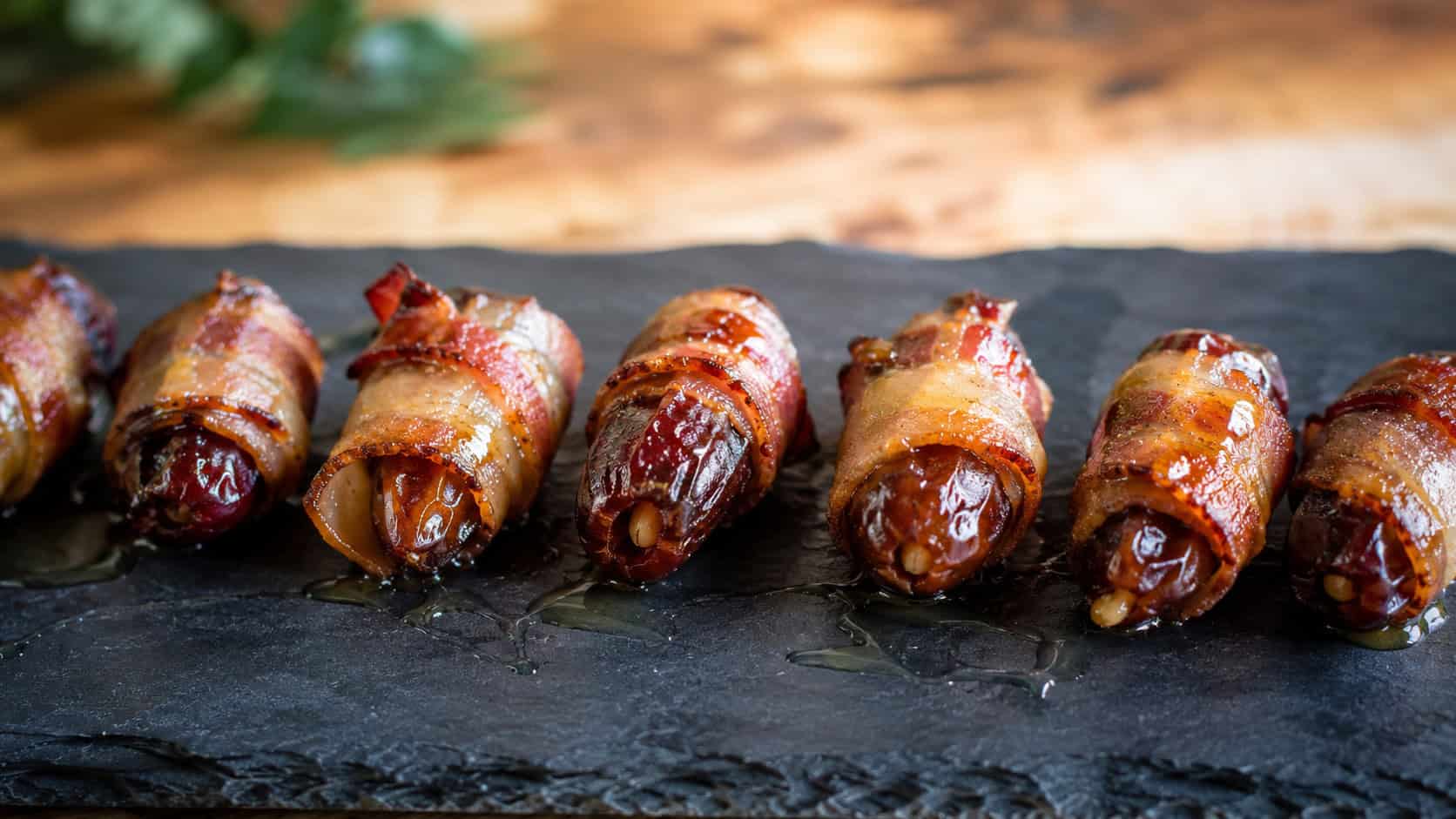 bacon-wrapped dates with crisp golden bacon drizzled with honey, arranged in a wide horizontal row on a slate board, glistening, warm natural light, shallow depth of field, landscape orientation