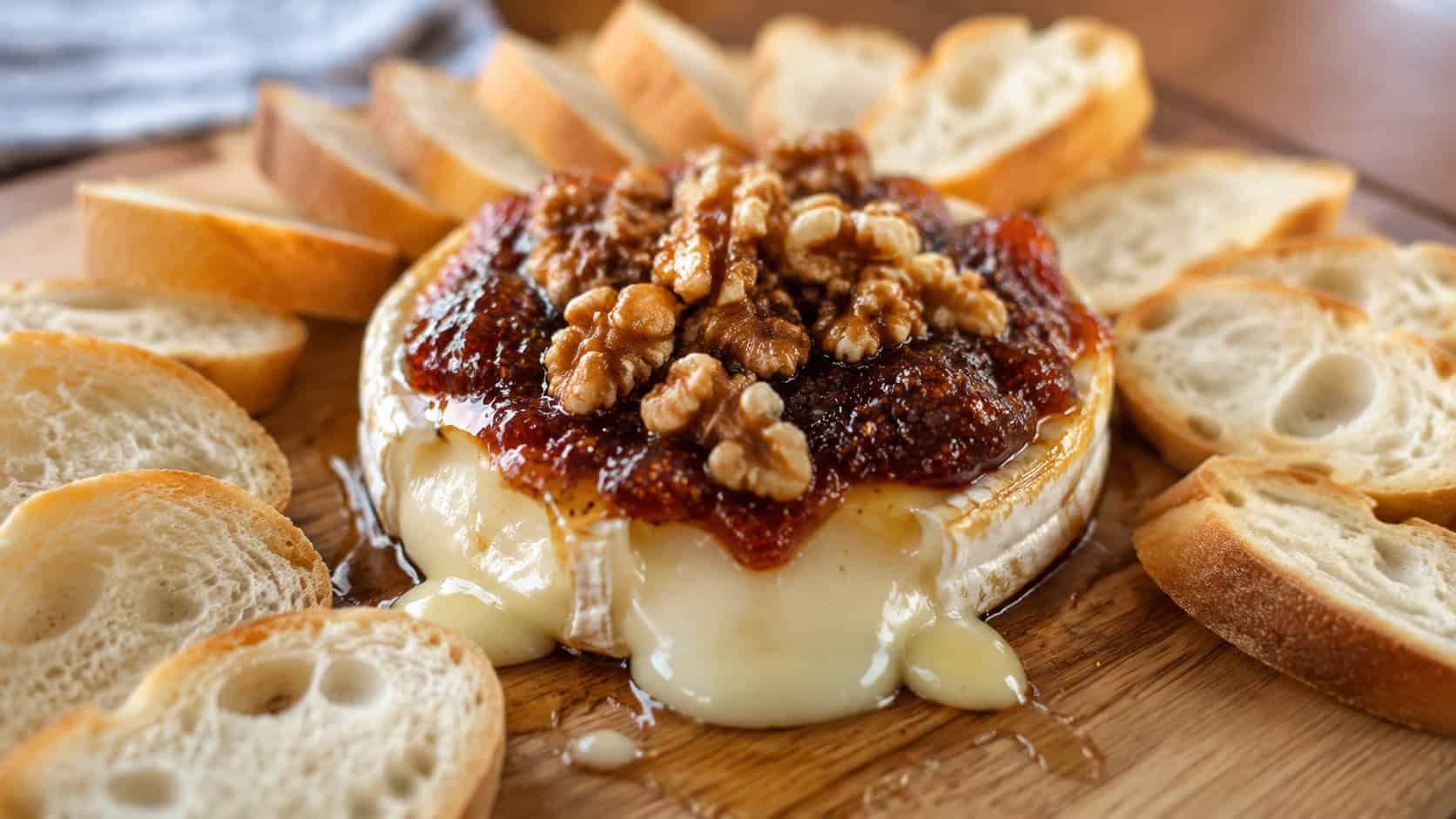 a molten baked brie topped with fig jam and walnuts drizzled with honey, in a wide horizontal shot on a wooden board surrounded by baguette slices, natural light, shallow depth of field, landscape orientation