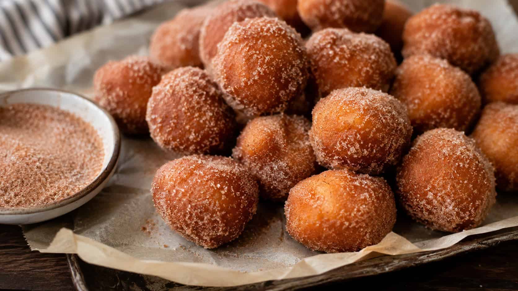 a pile of cinnamon sugar doughnut holes dusted golden brown, in a wide horizontal shot on a parchment-lined tray with a small bowl of cinnamon sugar, natural light, shallow depth of field, landscape orientation
